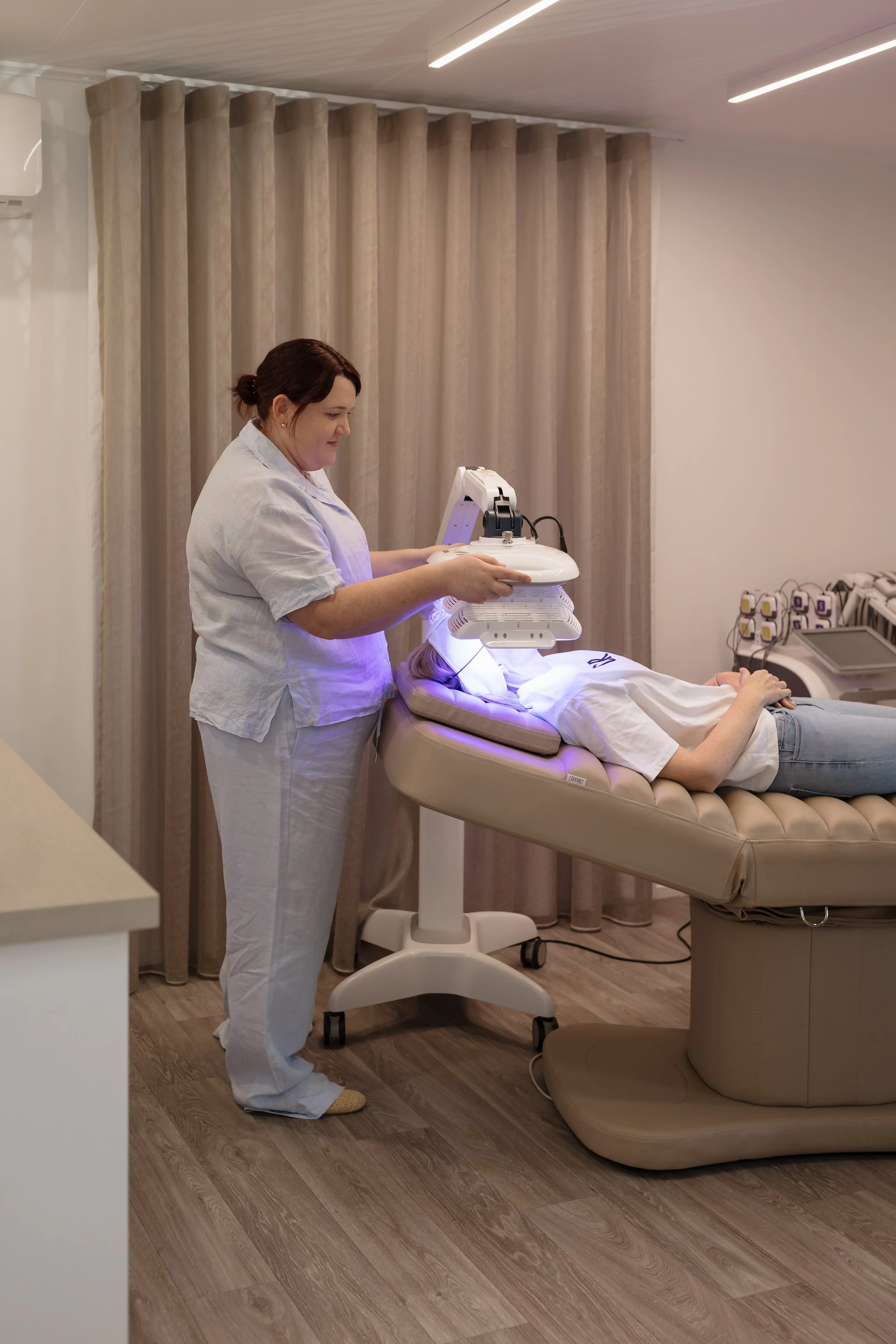 A woman lying on a medical bed receiving a treatment with a large machine overhead, while a medical professional operates the device in a clinical room with beige curtains and medical equipment.