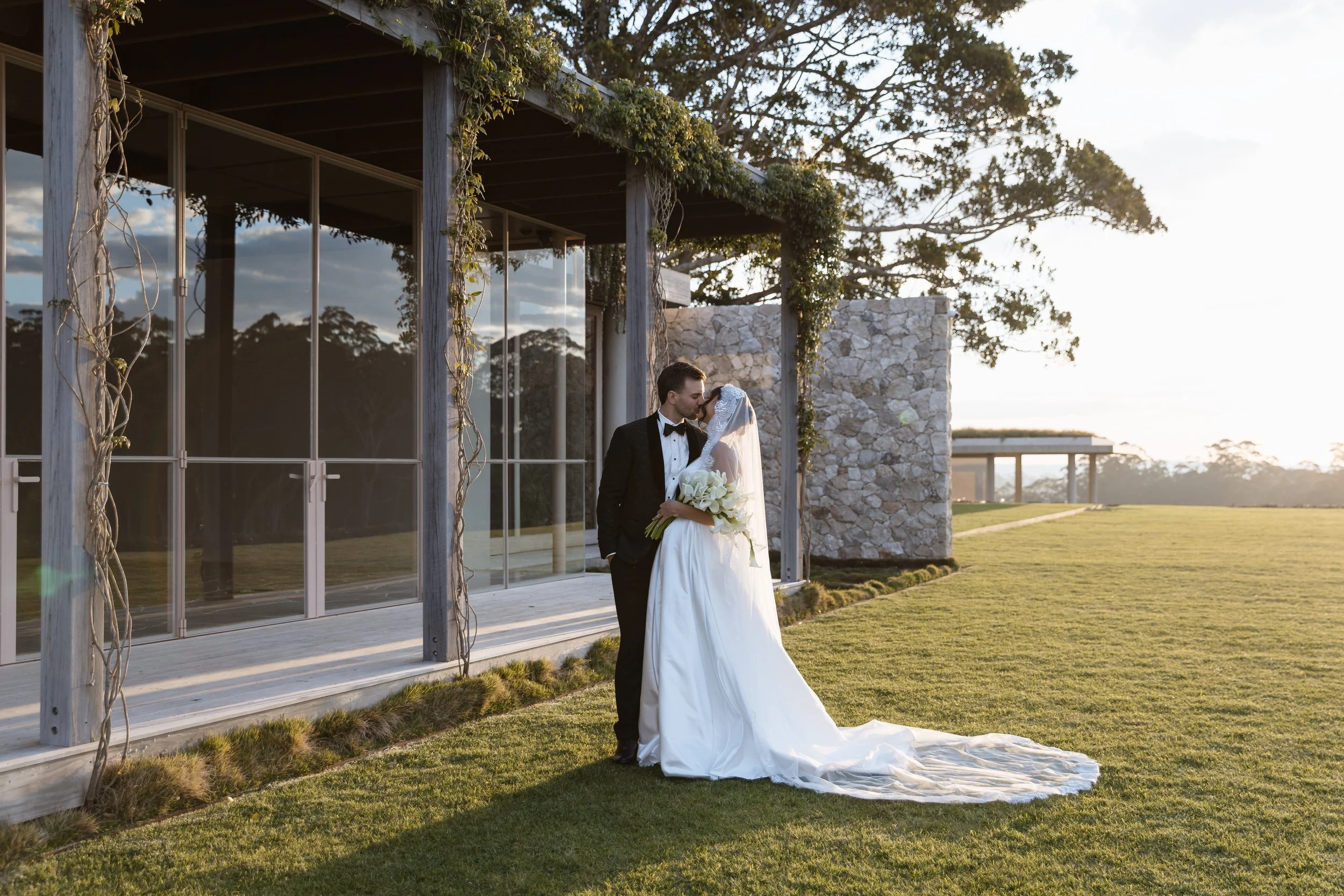 A bride and groom sharing a kiss outdoors during their wedding, with a modern building and trees in the background.