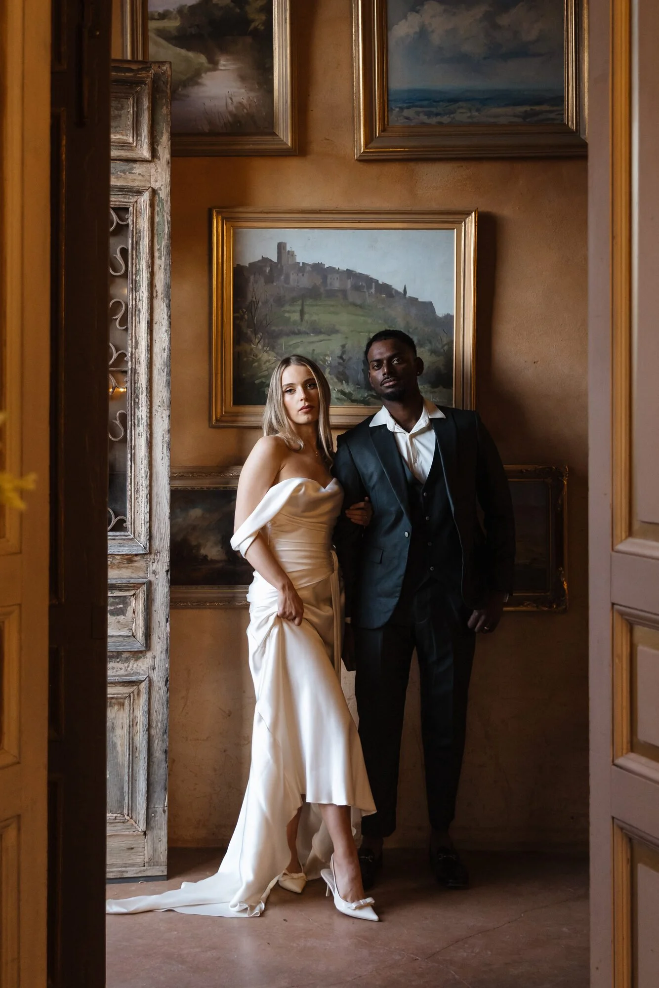 A man and woman stand together in a dimly lit room with several landscape paintings on the wall behind them. The woman wears a strapless white dress with a long train, high heels, and has light hair. The man wears a black suit with a white shirt and 