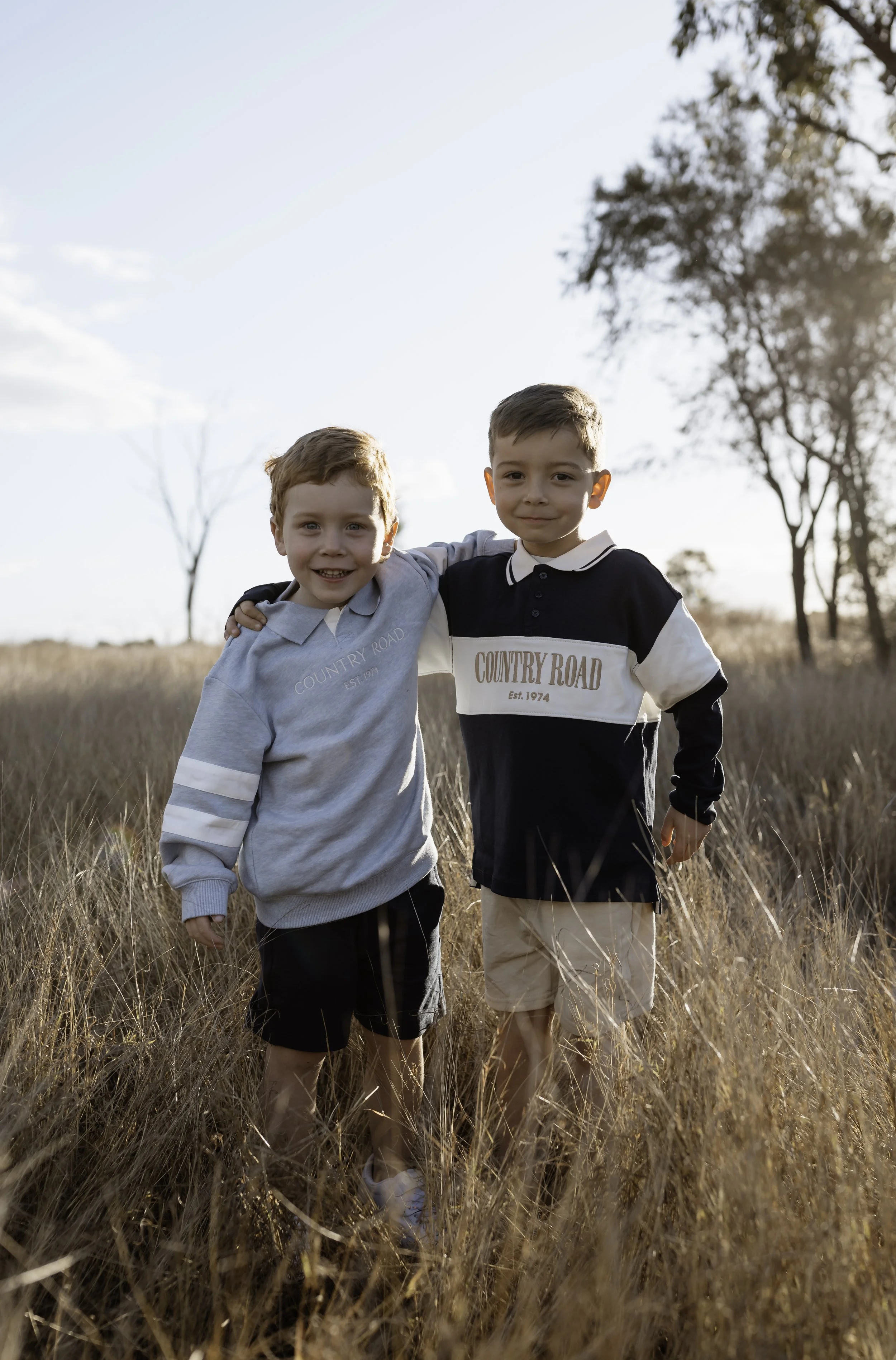 Two young boys smiling and standing closely together in a field of tall, dry grass during late afternoon or early evening sunlight. The background features a clear sky and trees.