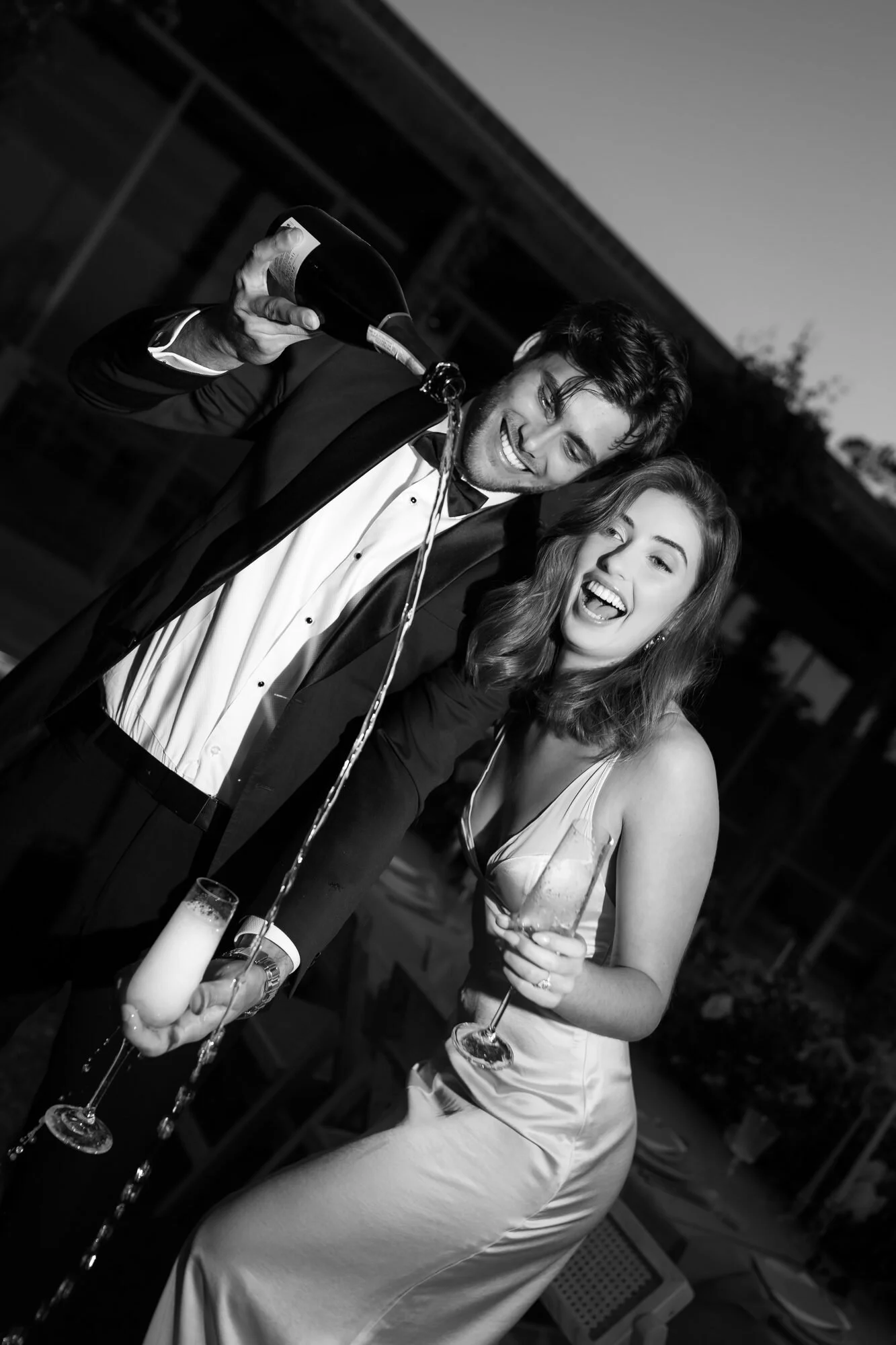 A young man in a tuxedo pours champagne into a fluted glass held by a young woman in a satin dress at an outdoor evening event.