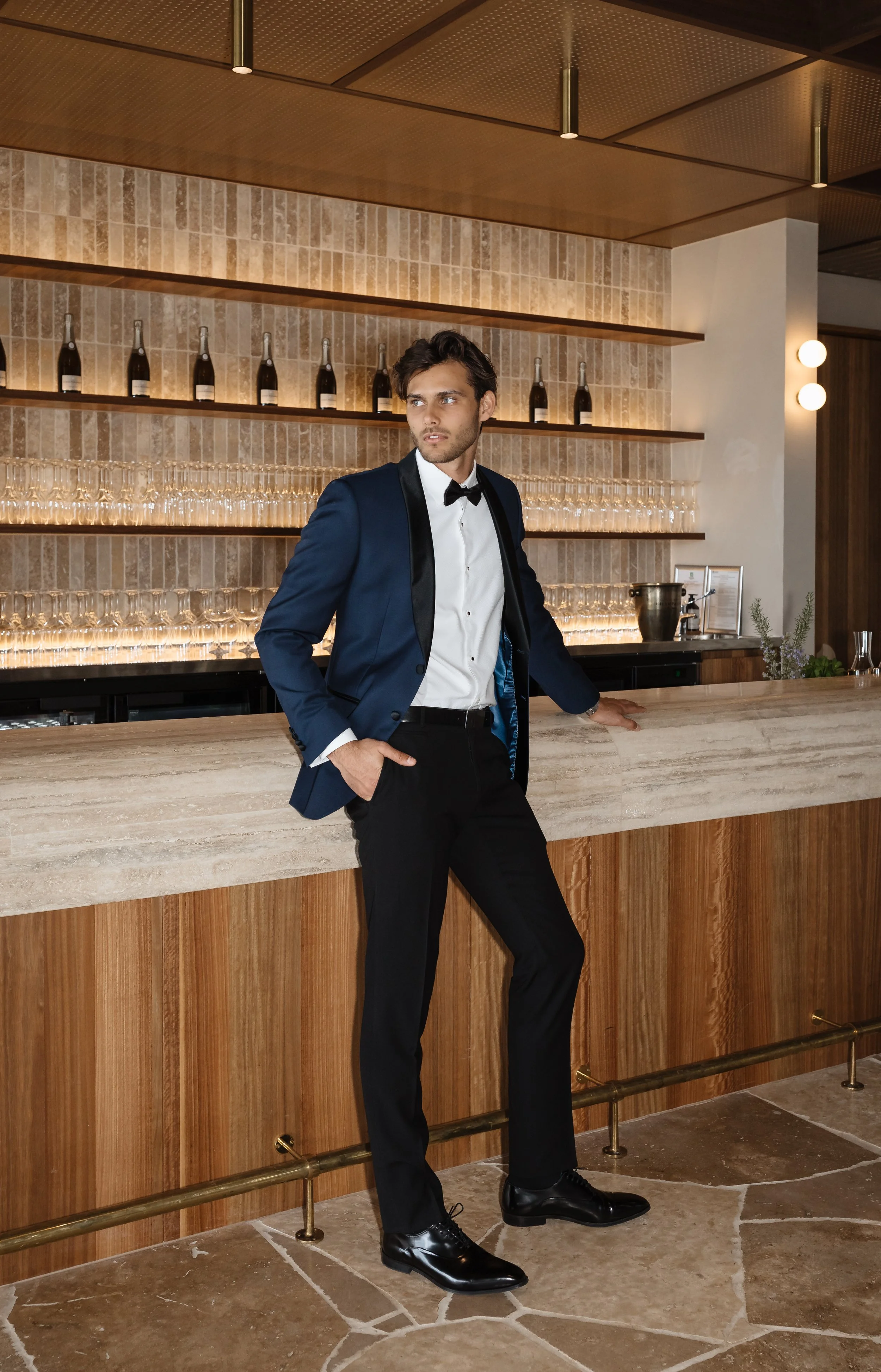 A young man in a tuxedo with a bowtie stands at a bar in a stylish restaurant or lounge. He is leaning against the bar with one hand in his pocket and looking to the side. Behind him are shelves with bottles of alcohol and a row of wine glasses. The 