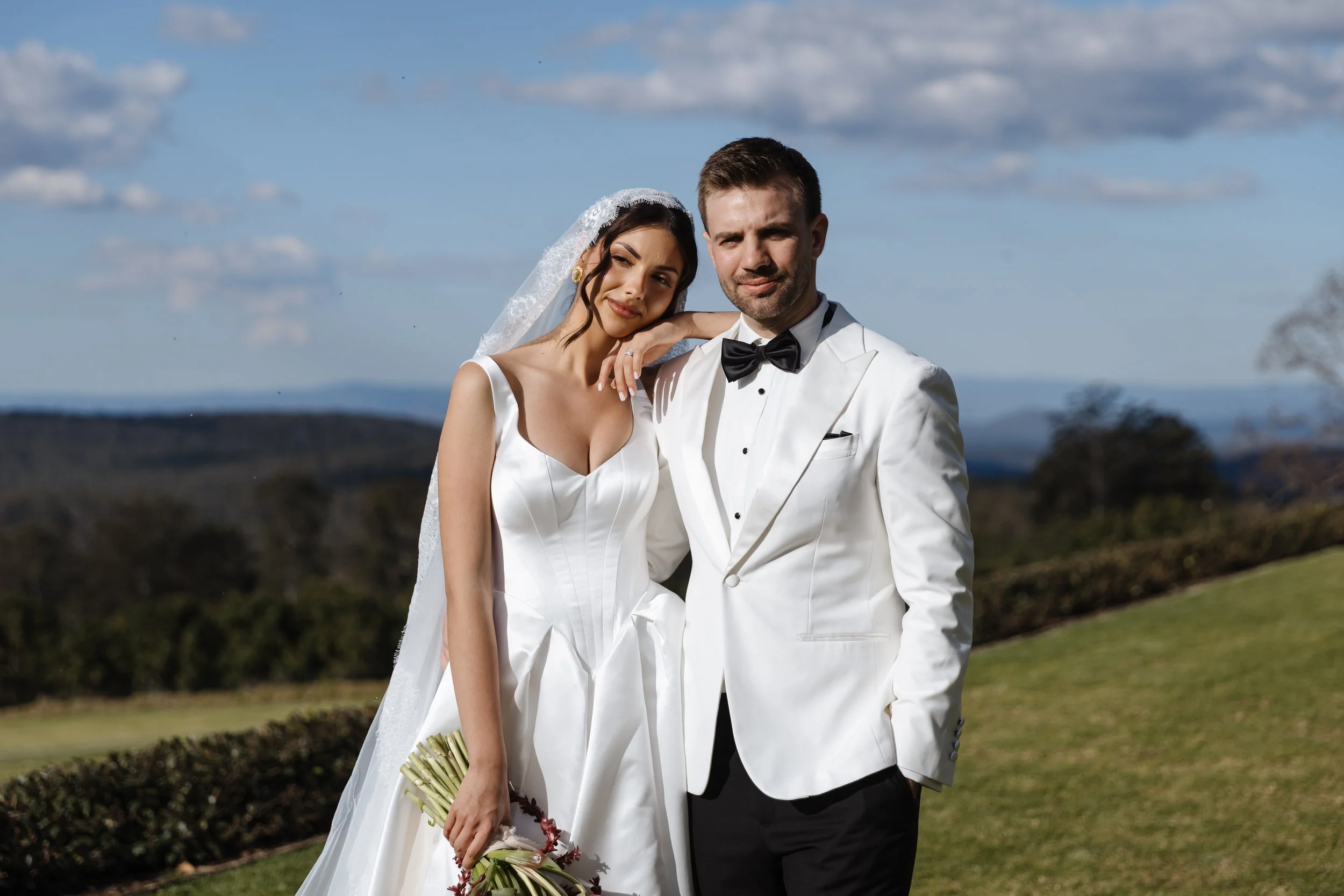 A bride and groom standing outdoors on their wedding day, with the bride holding a bouquet and the groom wearing a tuxedo, backdropped by a landscape with clouds, trees, and hills.