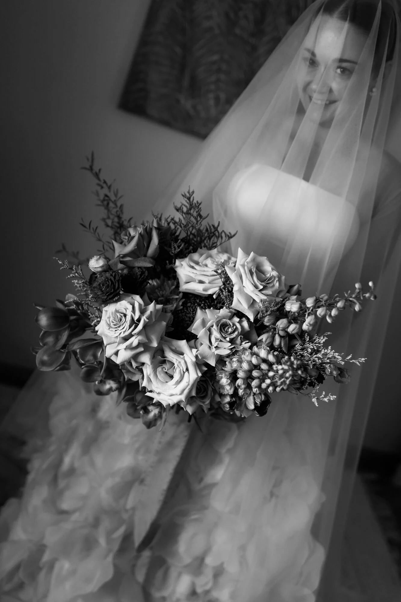 A woman wearing a veil holds a bouquet of roses and greenery, smiling.