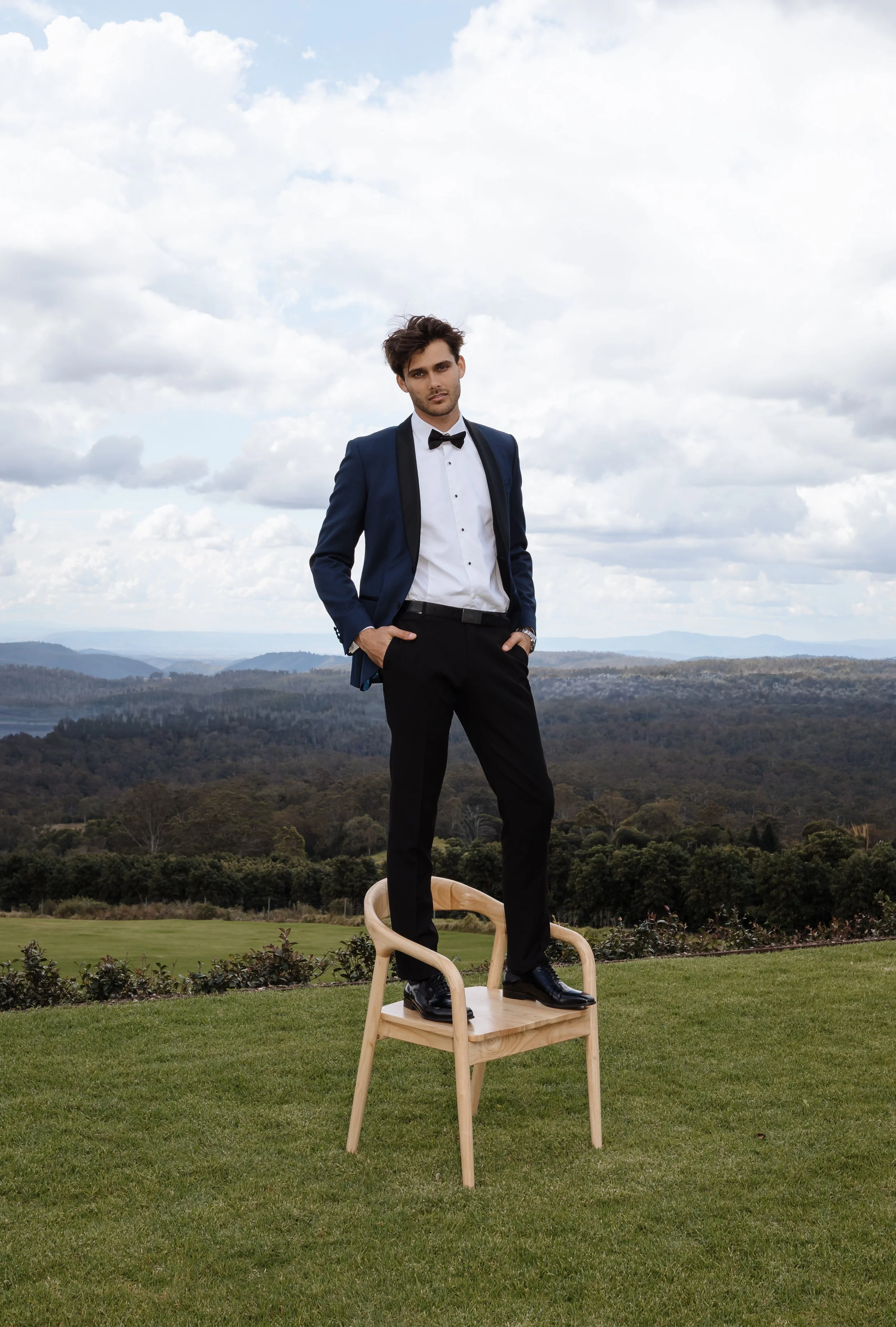 A young man in a tuxedo standing on a chair outdoors with a backdrop of hills and a cloudy sky.