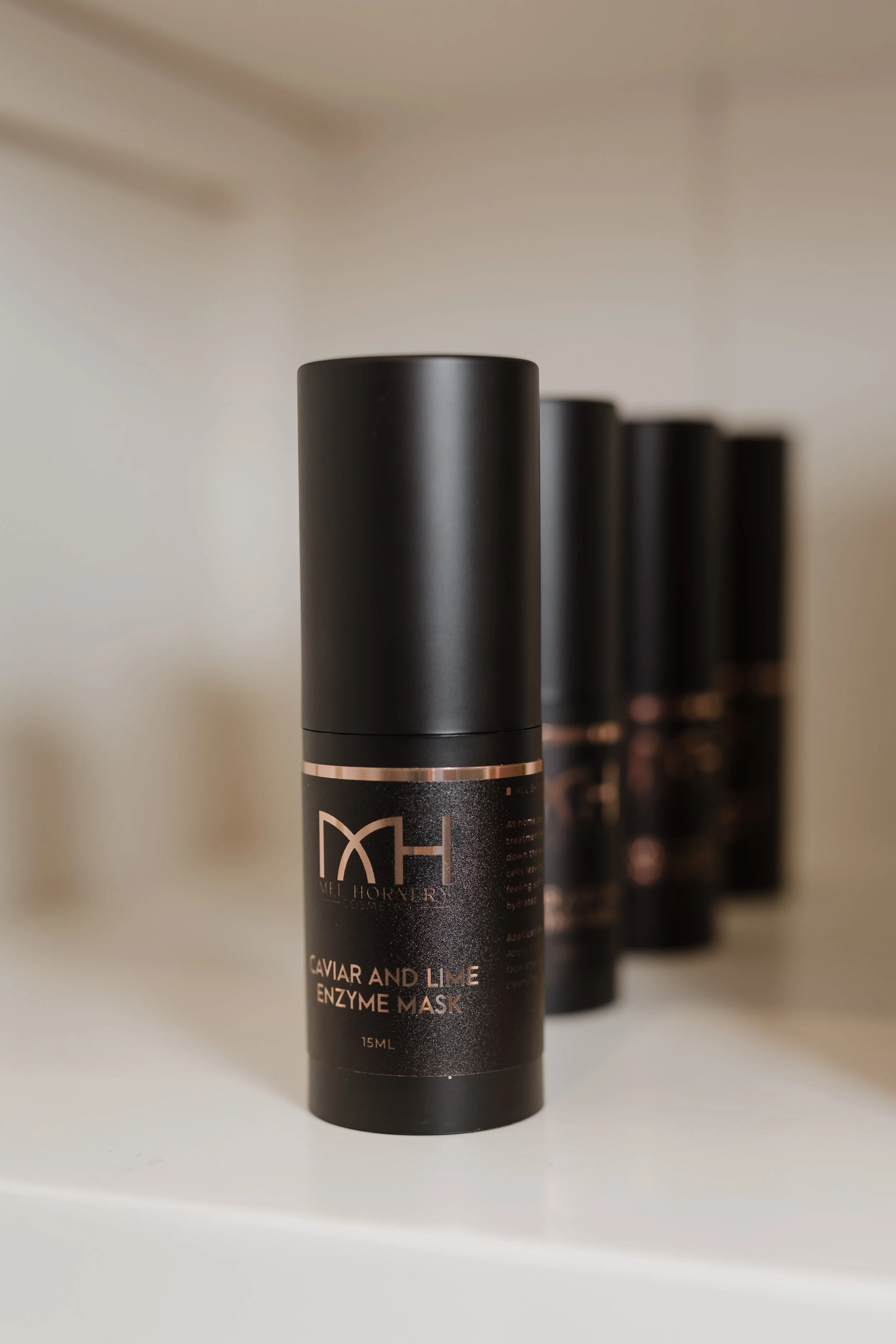 Black cylindrical bottles of skincare products lined up on a white surface, with the front one labeled 'Caviar and Lime Enzyme Mask' by Melhoney Cosmetics.