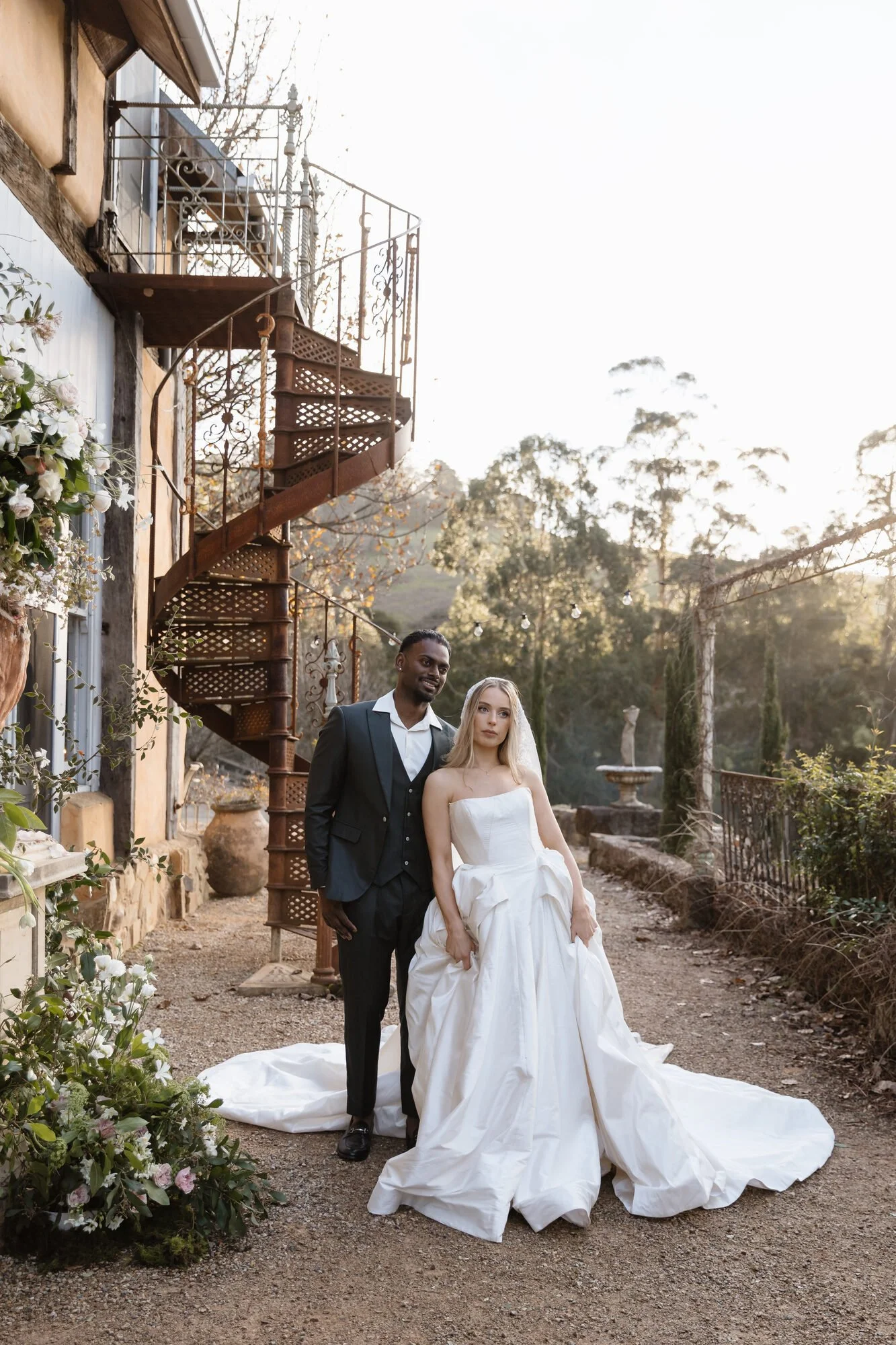 A bride and groom walking outdoors in a garden setting during sunset, with the bride in a strapless wedding gown and the groom in a dark suit.