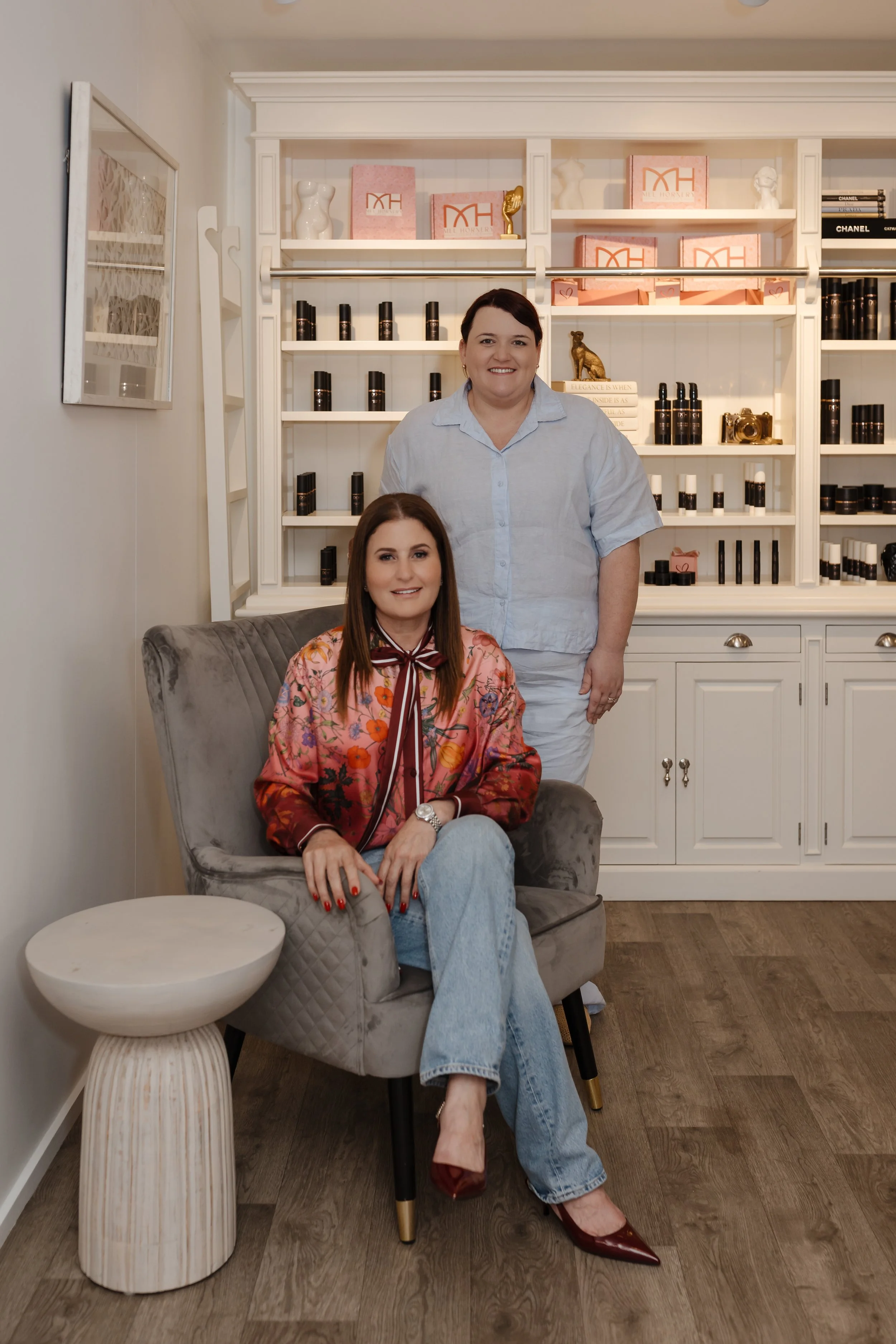 Two women pose in a room with a white bookshelf filled with decorative objects, books, and pink boxes. One woman is sitting on a gray velvet chair, wearing a floral jacket and light blue jeans, with red shoes. The other woman is standing behind her, 