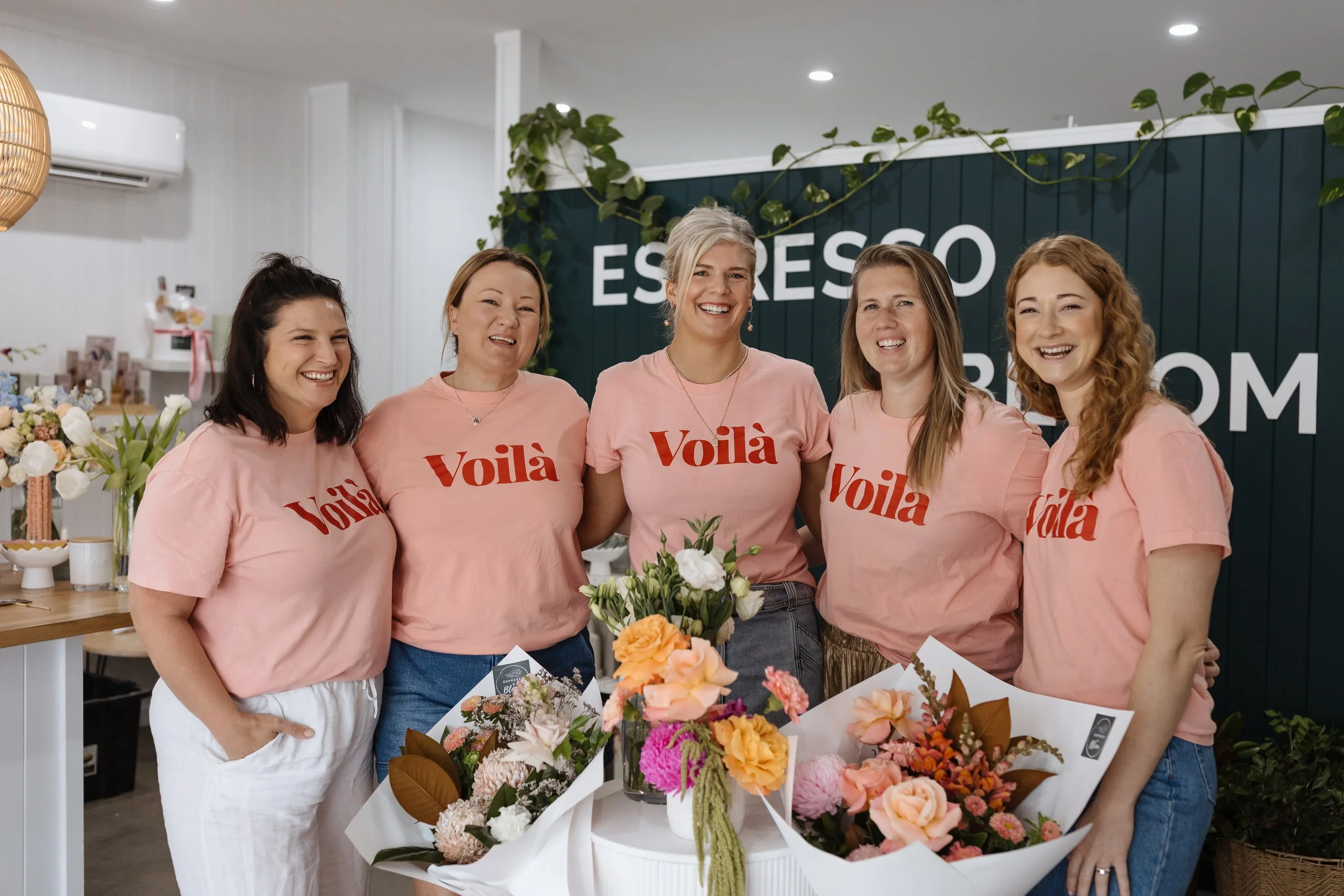 Group of five women smiling at a floral arrangement display in a bright, modern space with a dark blue sign reading 'ESPREZZO' in the background. All women are wearing matching pink T-shirts with 'Voilà' written on them.