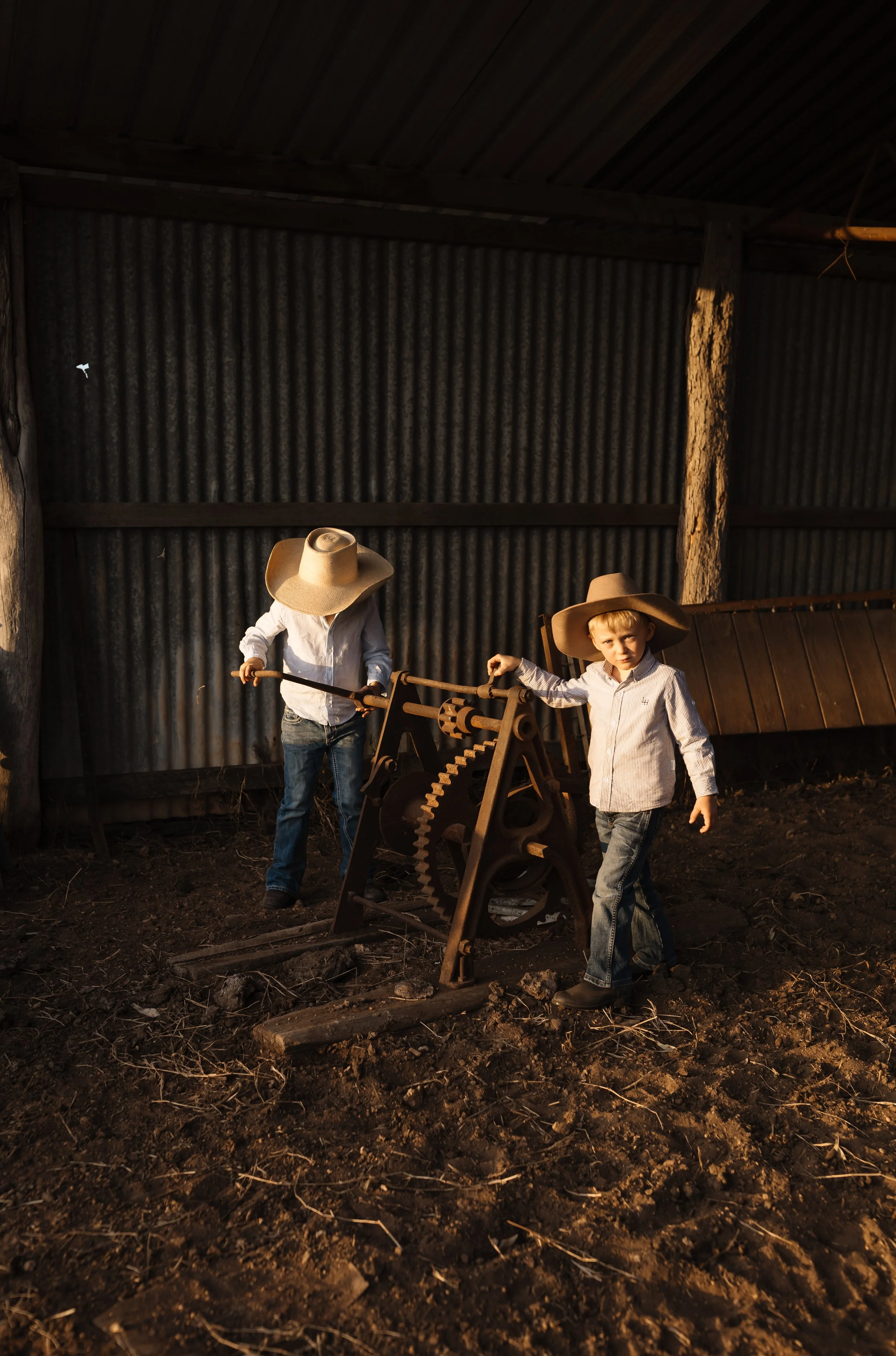 Two boys wearing large straw hats and casual shirts, standing outdoors in a rustic barn-like setting, handling vintage equipment on uneven, earthy ground.