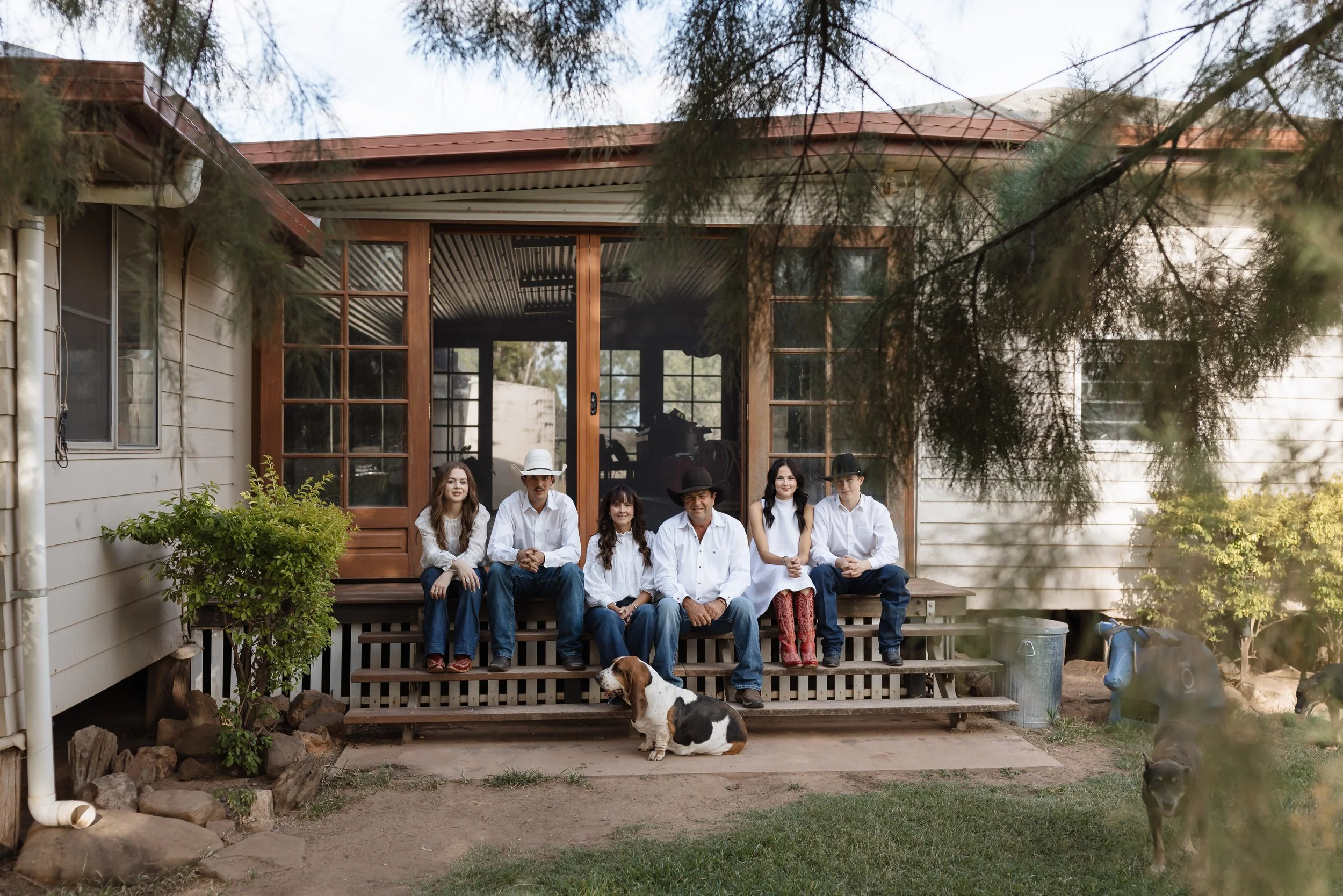 A family of seven posing on the front porch of a house, with a dog sitting in front. The family members are sitting and standing on the porch steps, with trees and the house visible in the background.