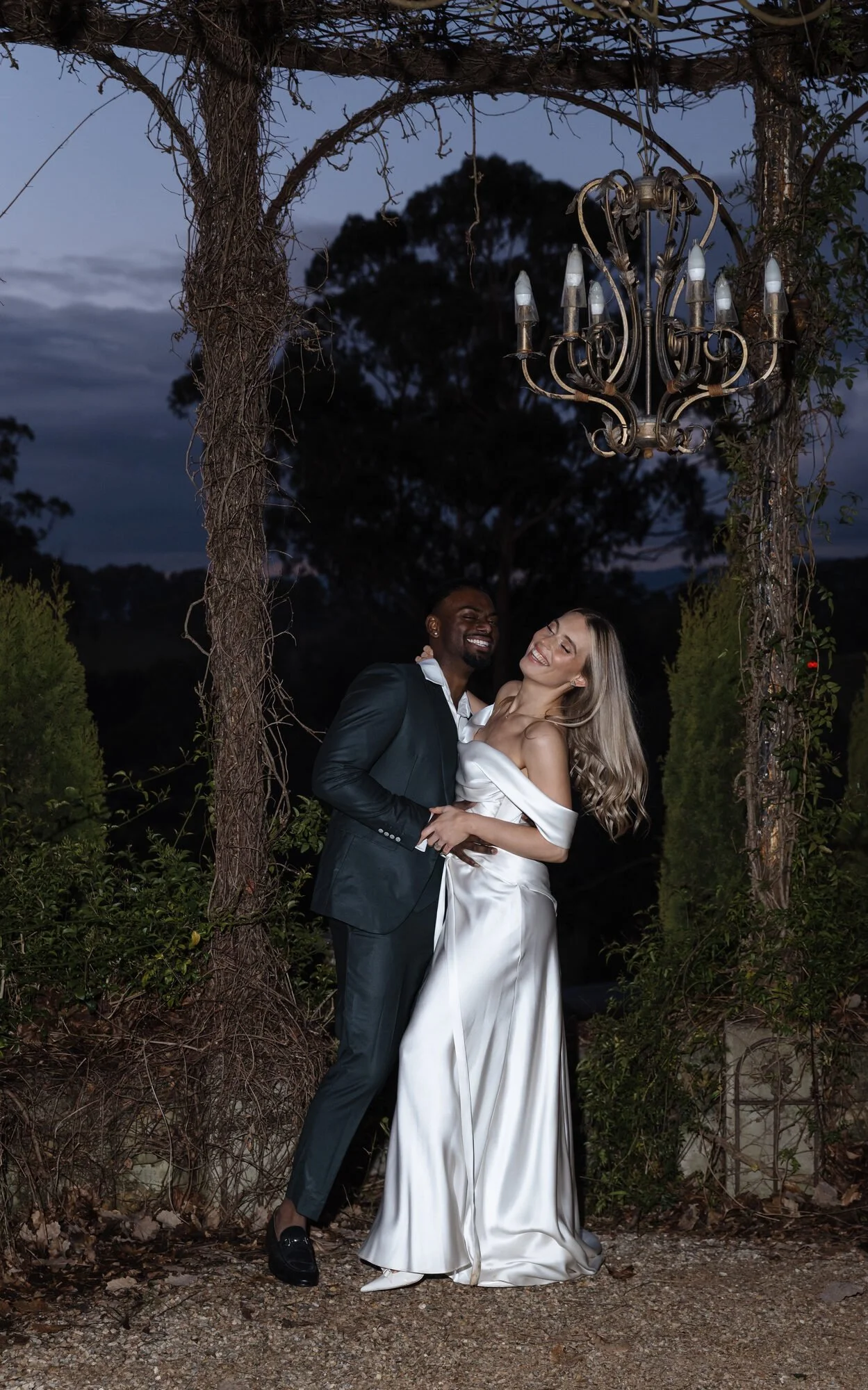 A couple in formal attire dancing outdoors at dusk under a chandelier hanging from a wooden arbor, surrounded by greenery.