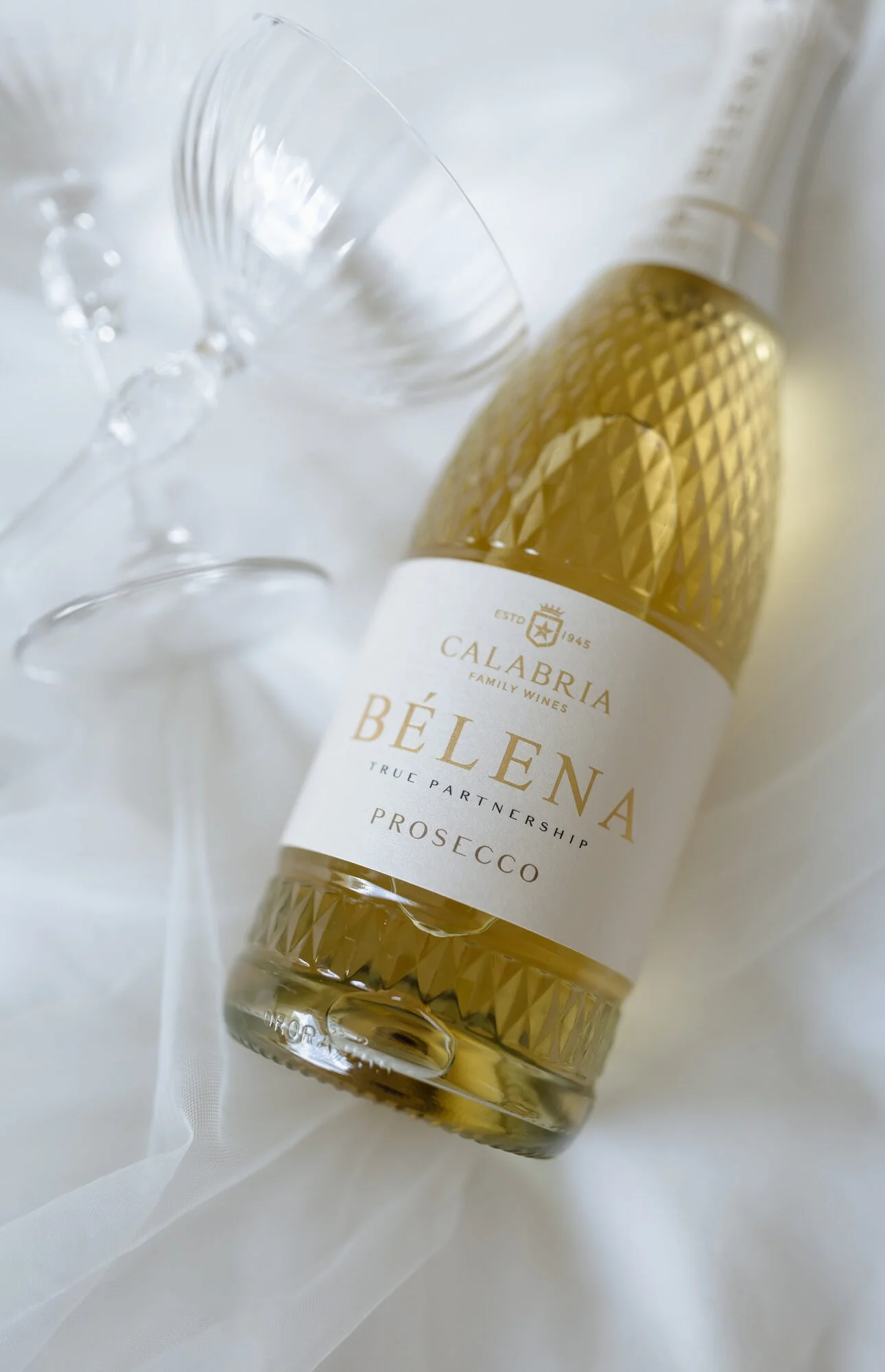 A bottle of Calarbria Belen Prosecco with a white label and gold accents, surrounded by a clear wine glass and a white fabric background.