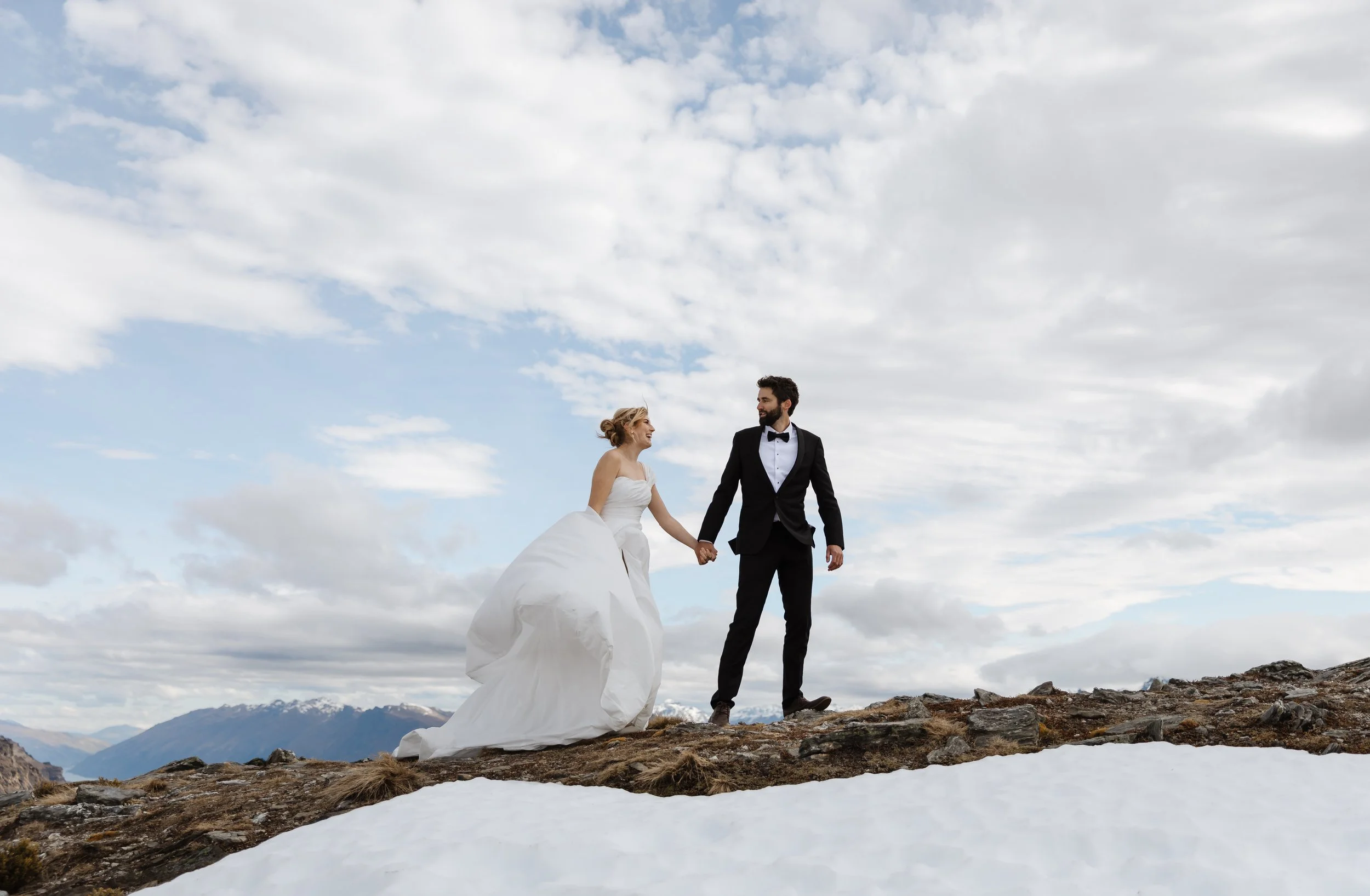 A bride and groom holding hands and walking on a rocky, snowy landscape under a cloudy sky, with mountains in the background.