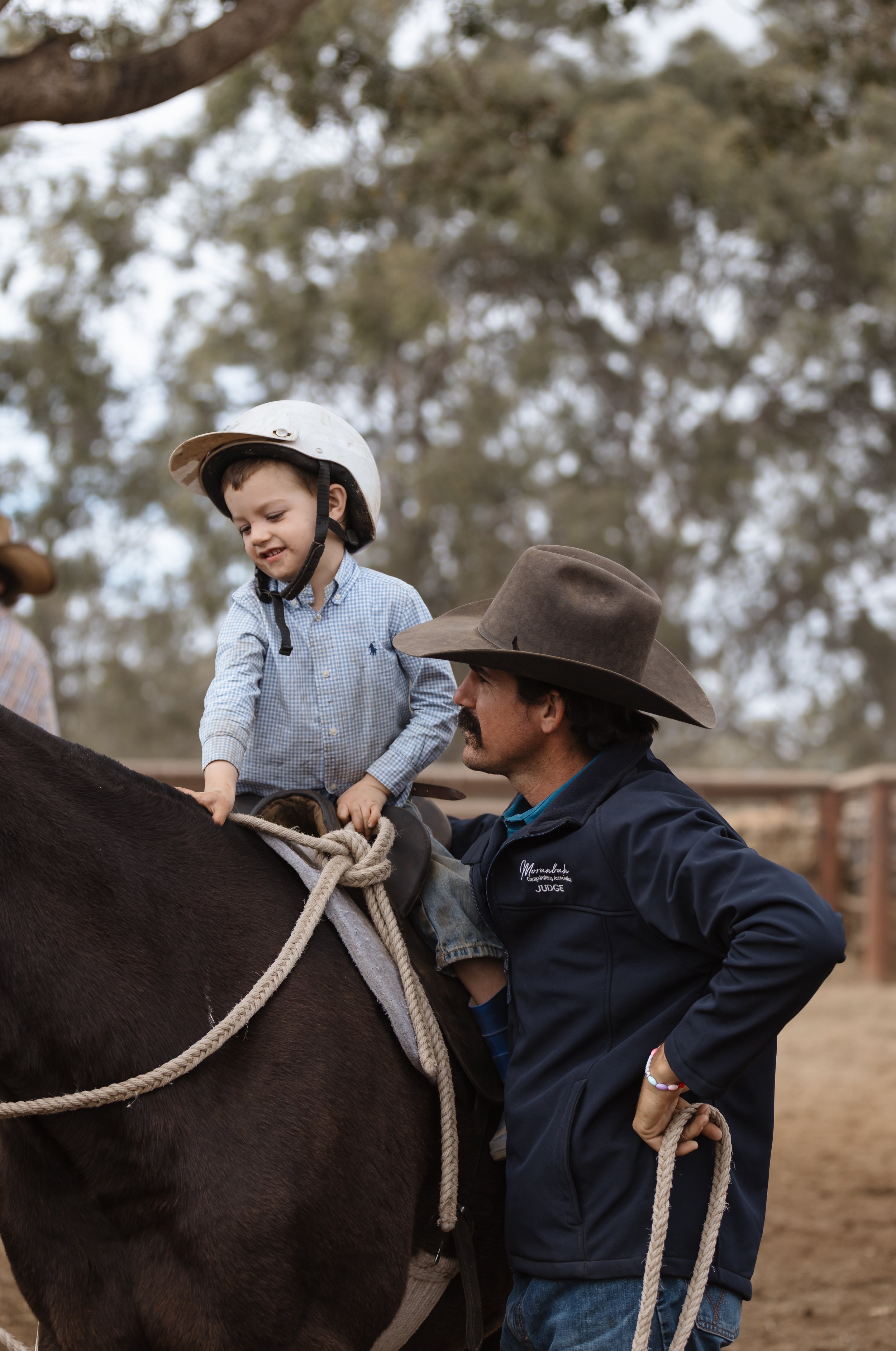 A young boy with a helmet riding on a horse, assisted by a man in a judge's jacket and a brown cowboy hat, outdoors with trees in the background.
