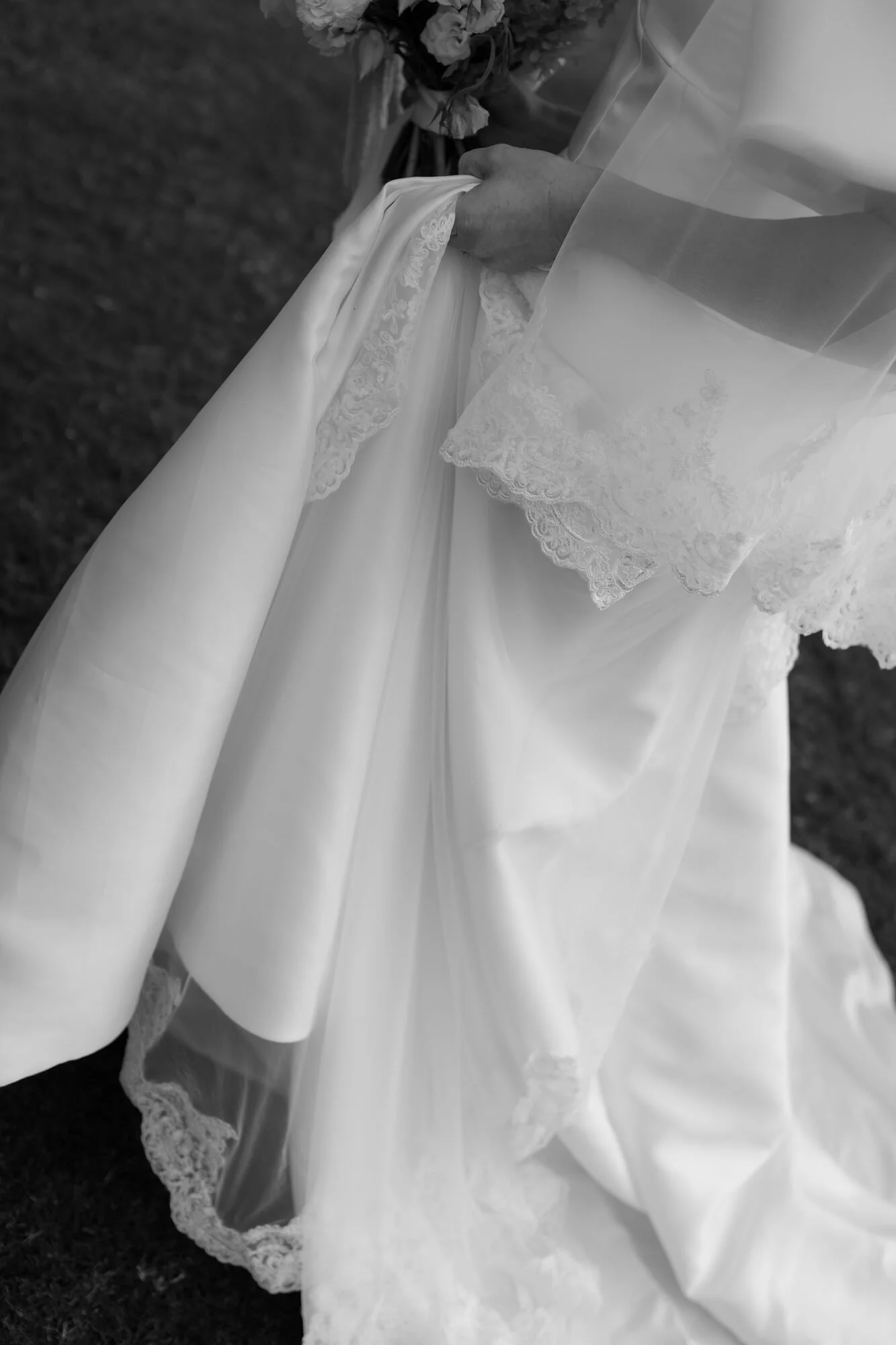 Close-up of a bride holding the hem of her wedding dress with lace details, outdoors on grass.