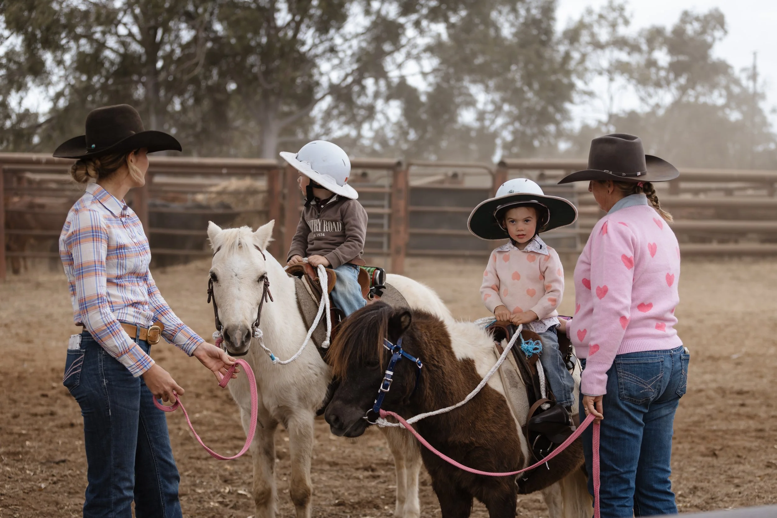 Two young girls sitting on ponies, wearing hats and looking at their instructor, with the instructor holding a pony's lead rope in a dirt riding area.