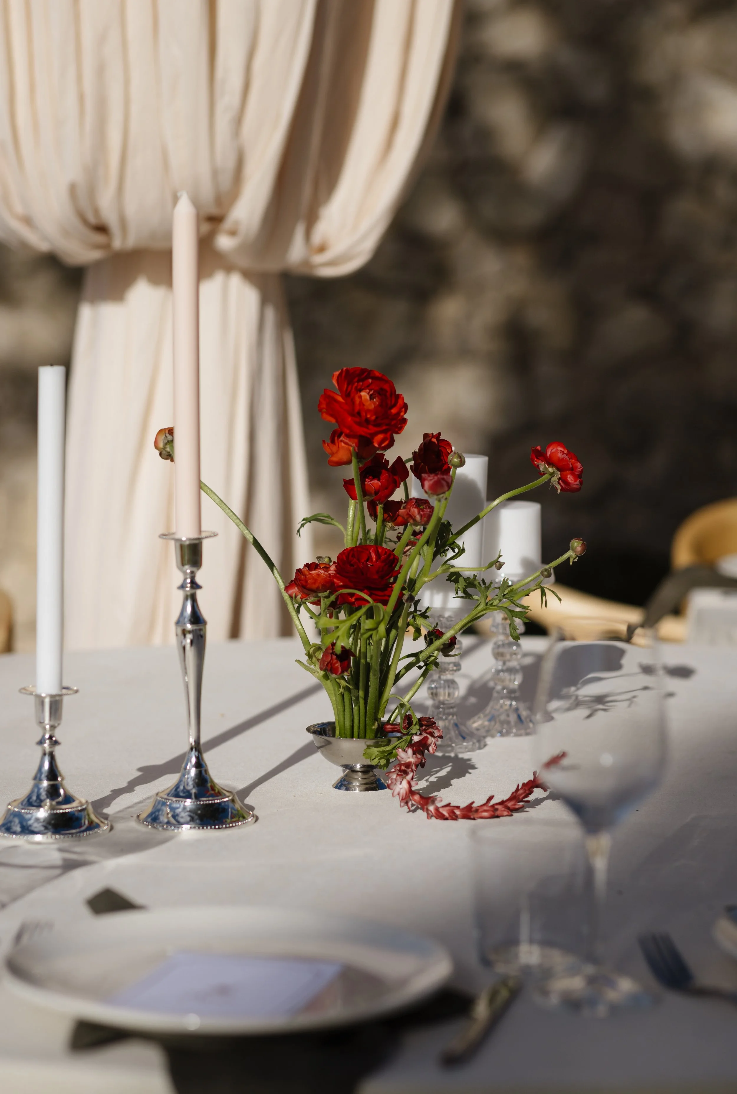 A table centerpiece with red flowers in a silver vase, surrounded by white candles, set on a white tablecloth, with a blurred stone wall background.