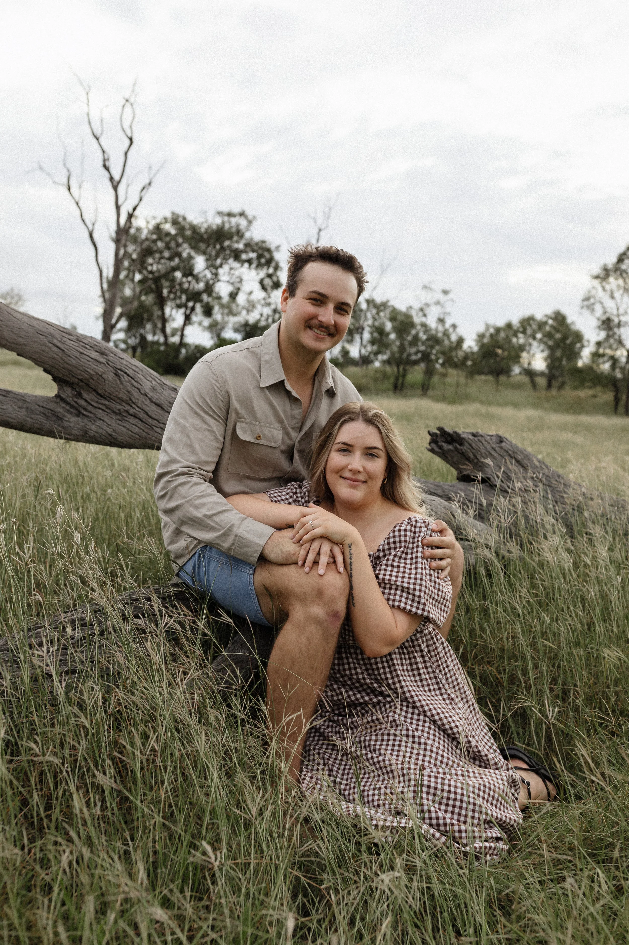 A young man and woman sitting together in a grassy field, with trees and cloudy sky in the background.