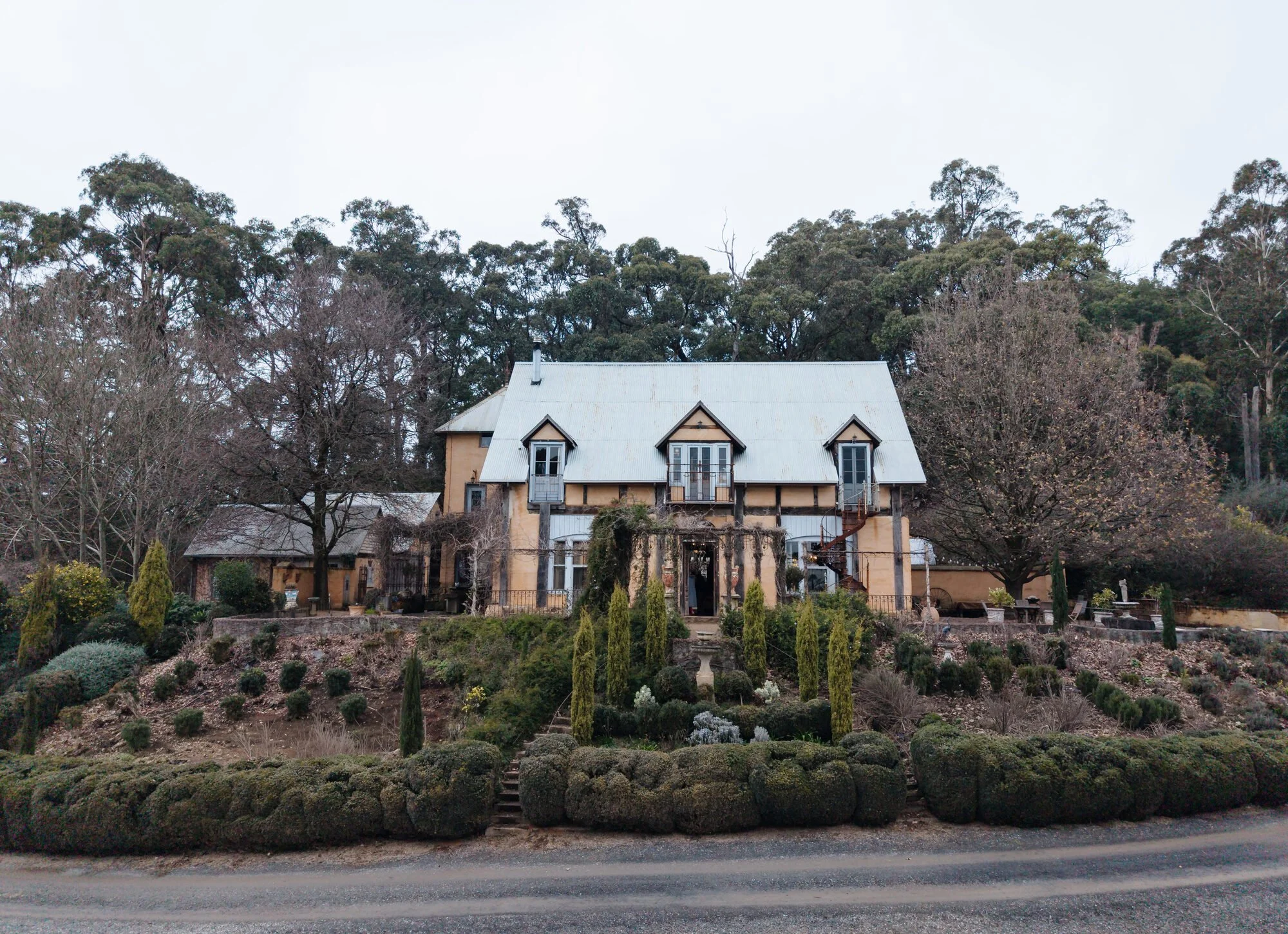 A house with a gray metal roof and wooden construction, surrounded by a landscaped garden with bushes, small trees, and stone steps leading up to the house, with bare trees and forested hillside in the background.