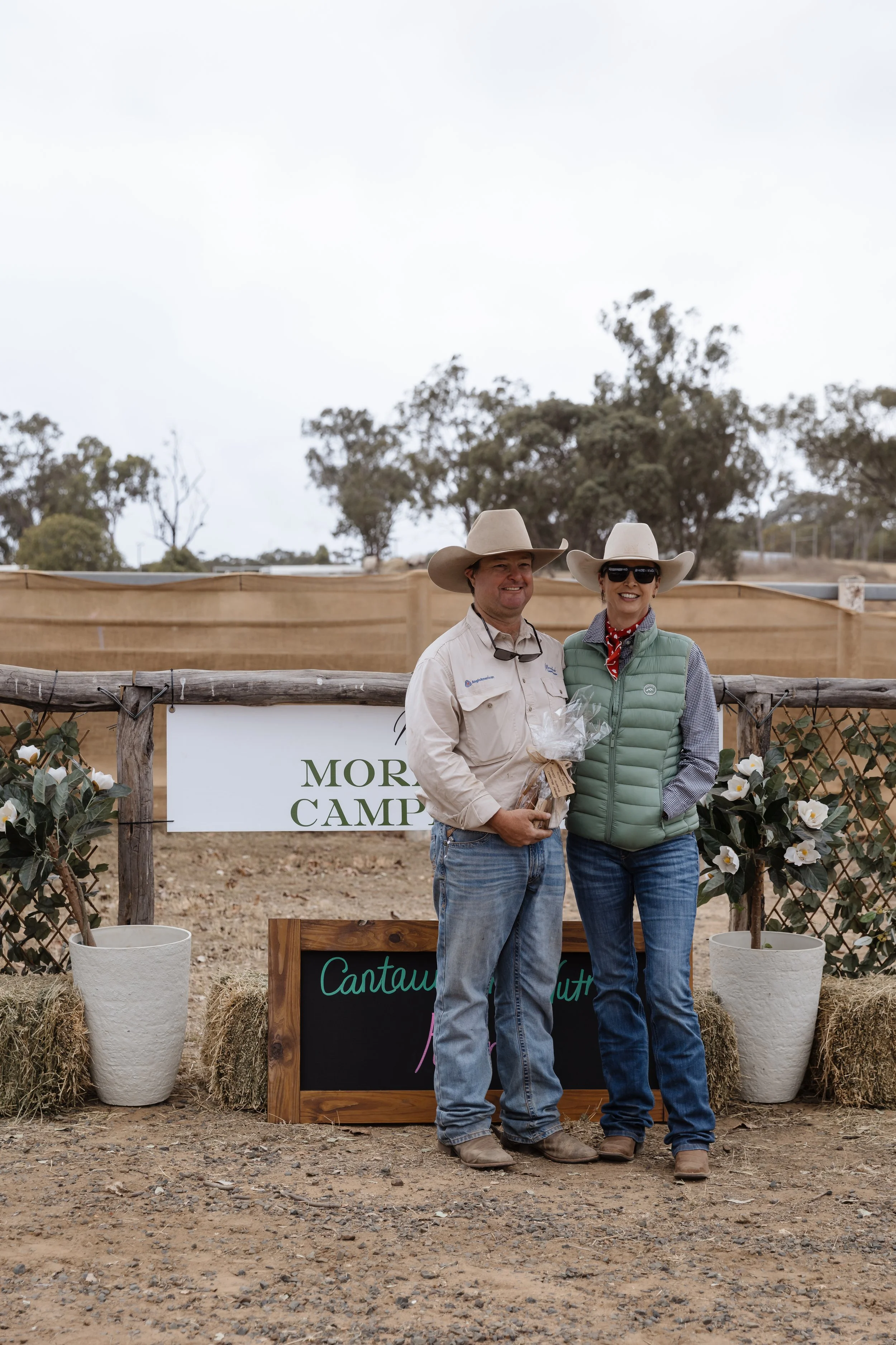 Two smiling people, a man and a woman, wearing cowboy hats standing outdoors in front of a rustic wooden fence with greenery and trees in the background. They are at an event with signs and flowers, and one is holding a gift. The ground is dirt.