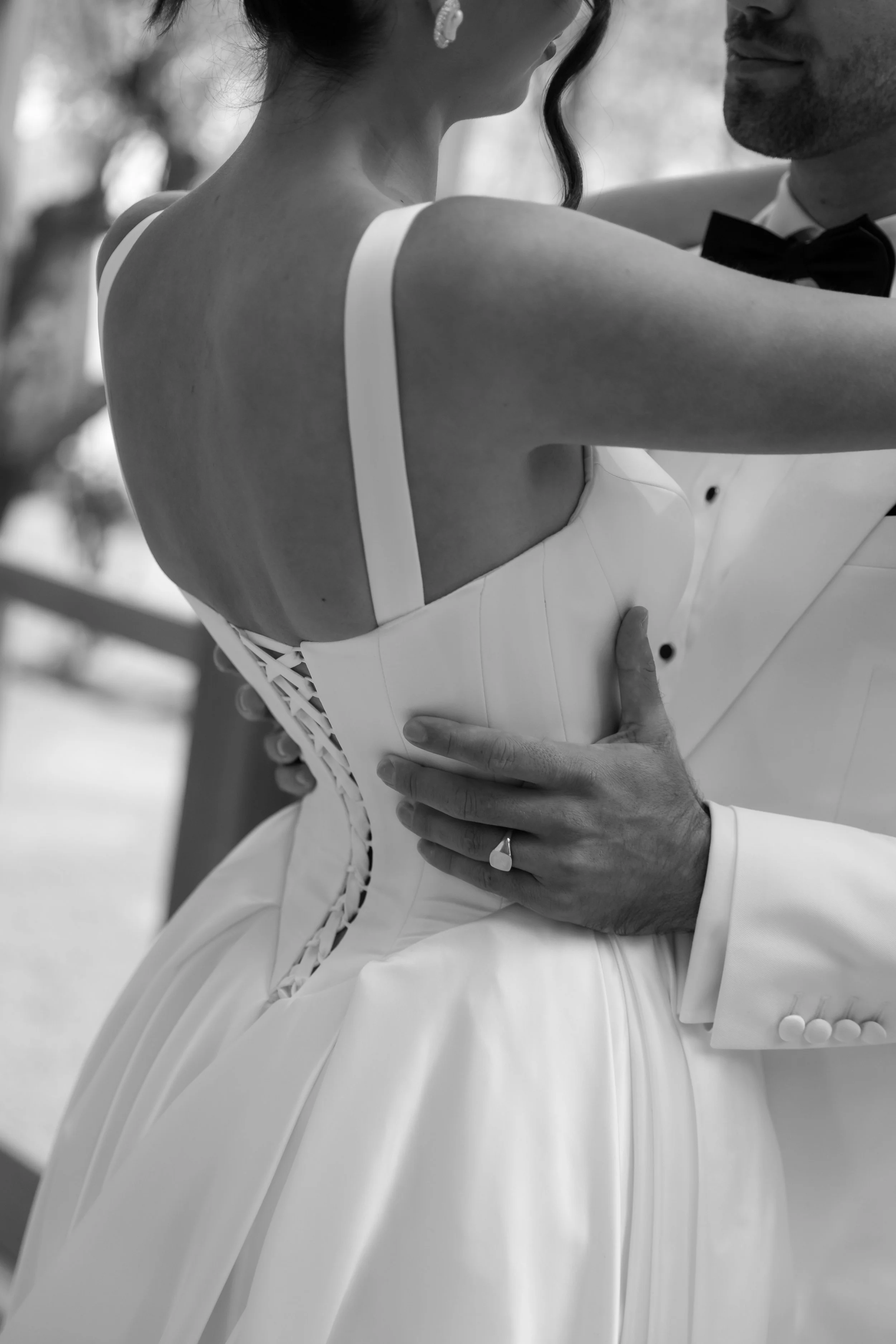 A black-and-white photo of a bride and groom dancing close together, with the bride in a sleeveless dress and the groom in a tuxedo. Their faces are not visible.