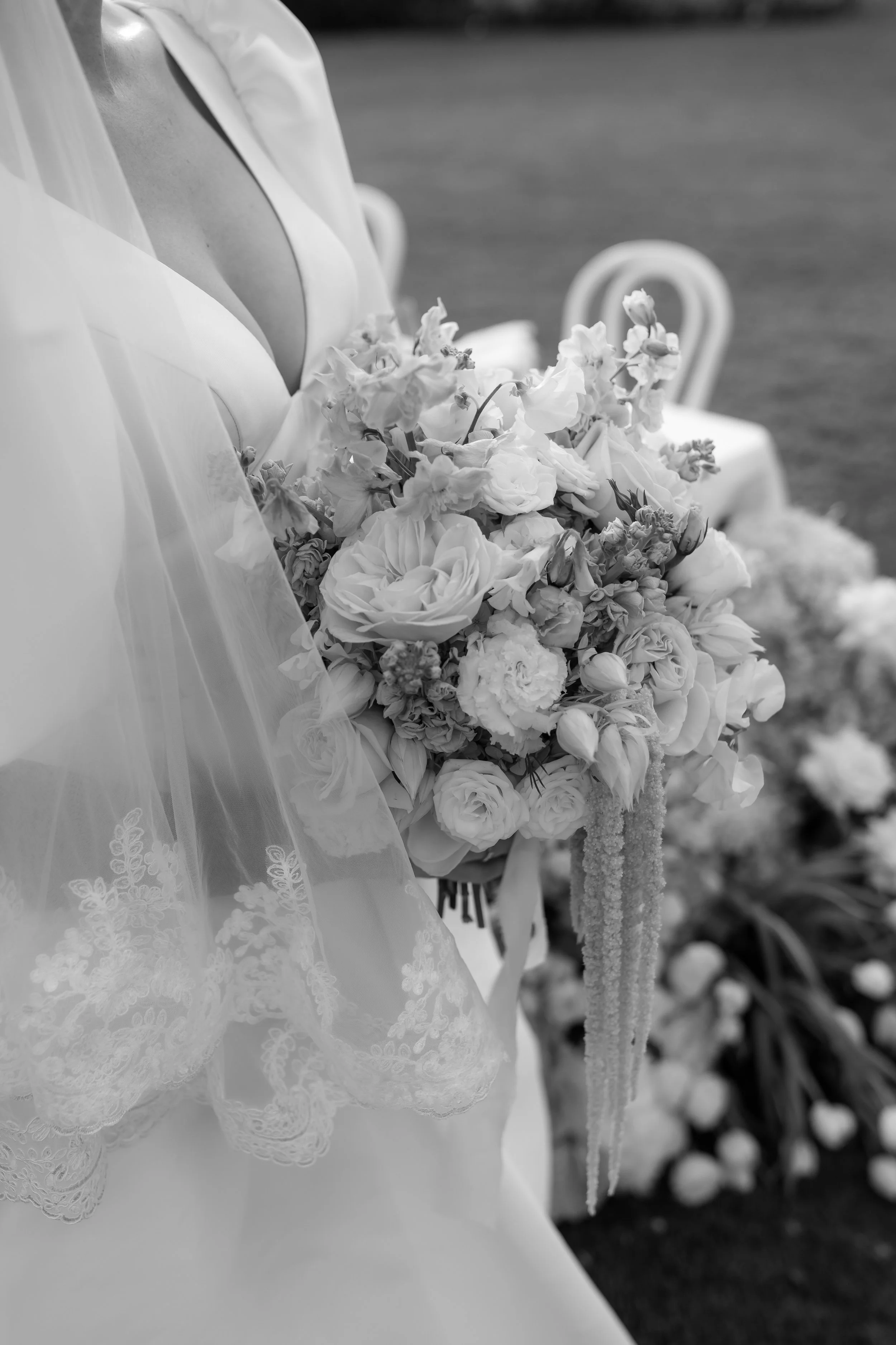 Black and white photo of a bride holding a bouquet of flowers, with a lace veil draped over her wedding dress, in an outdoor setting with chairs and greenery in the background.