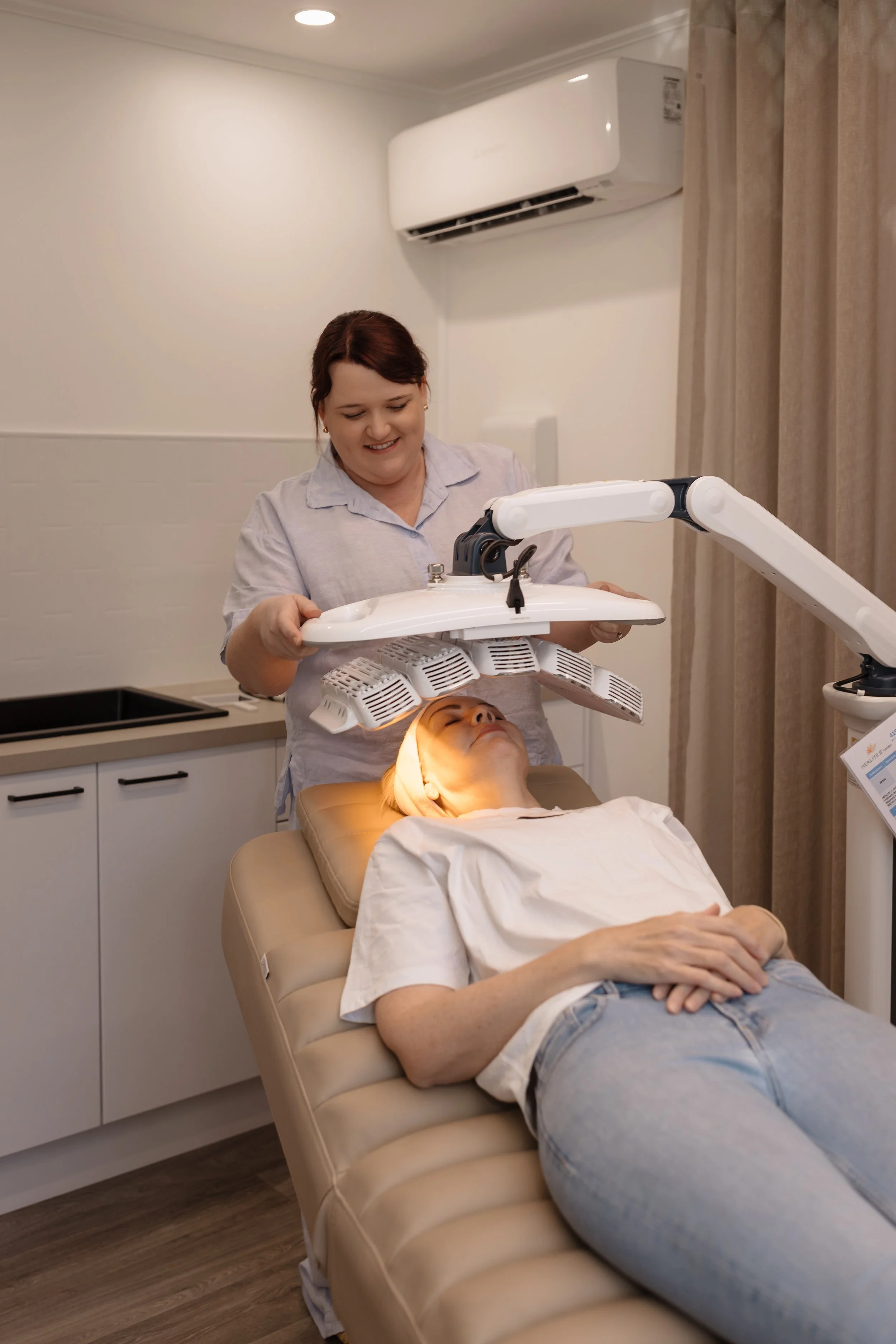 A woman with reddish-brown hair receiving a facial treatment or facial therapy in a spa or skincare clinic. She is lying on a beige treatment bed with her eyes closed and a relaxed expression.