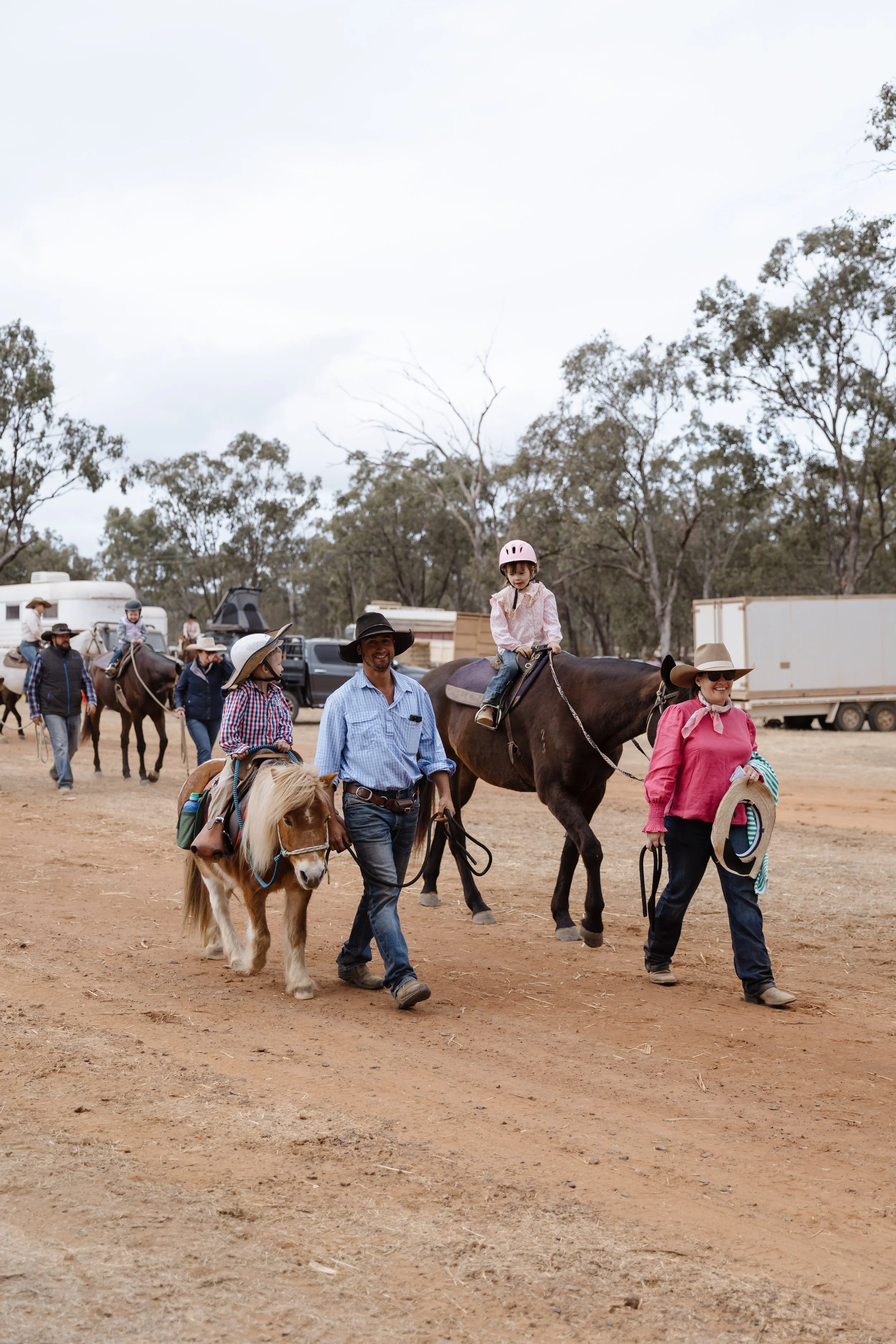 People riding and walking horses in a dirt outdoor area with trees and camping trailers in the background.