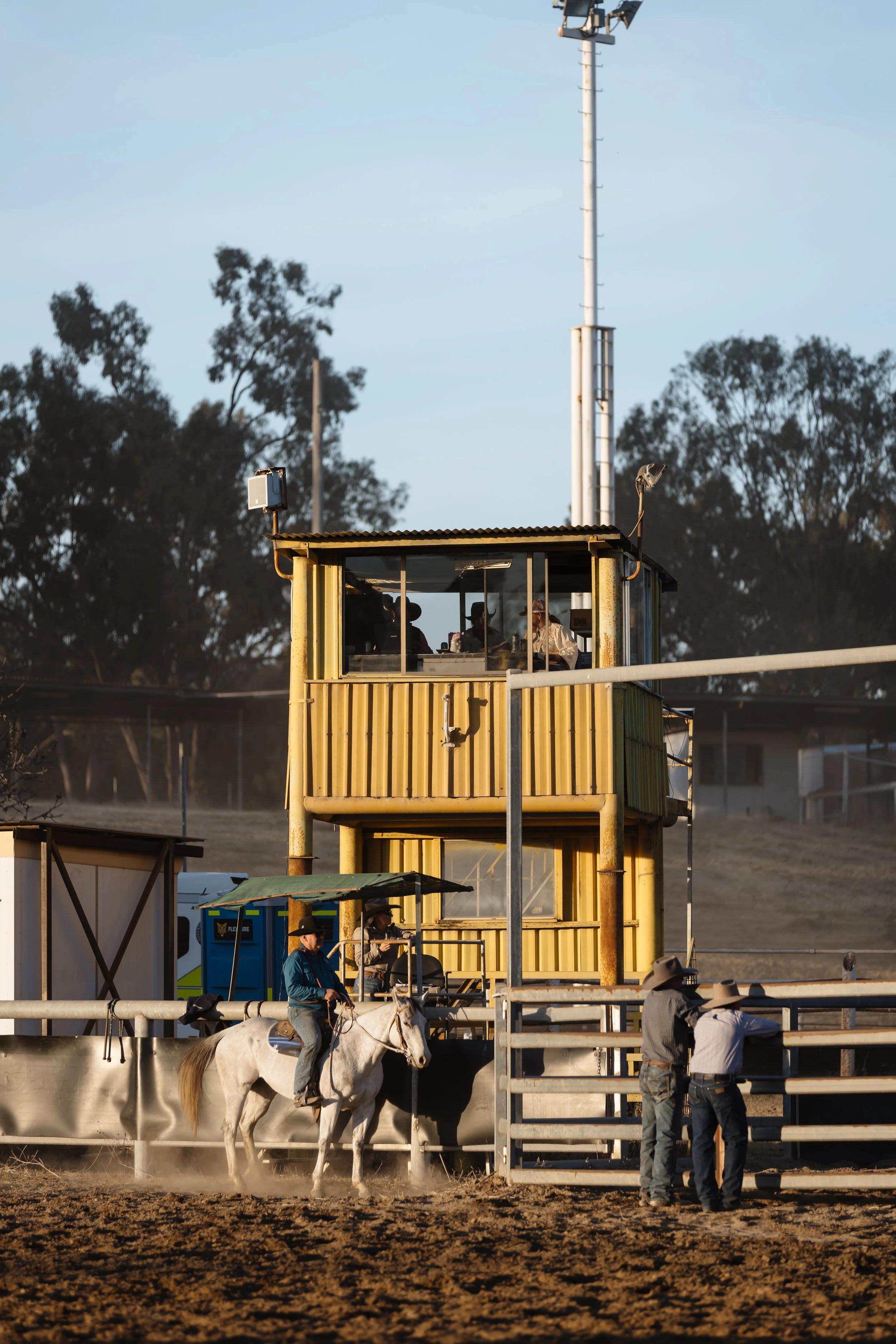 Riders on horseback approaching a yellow structure at a horse racing track, with officials nearby and trees in the background.