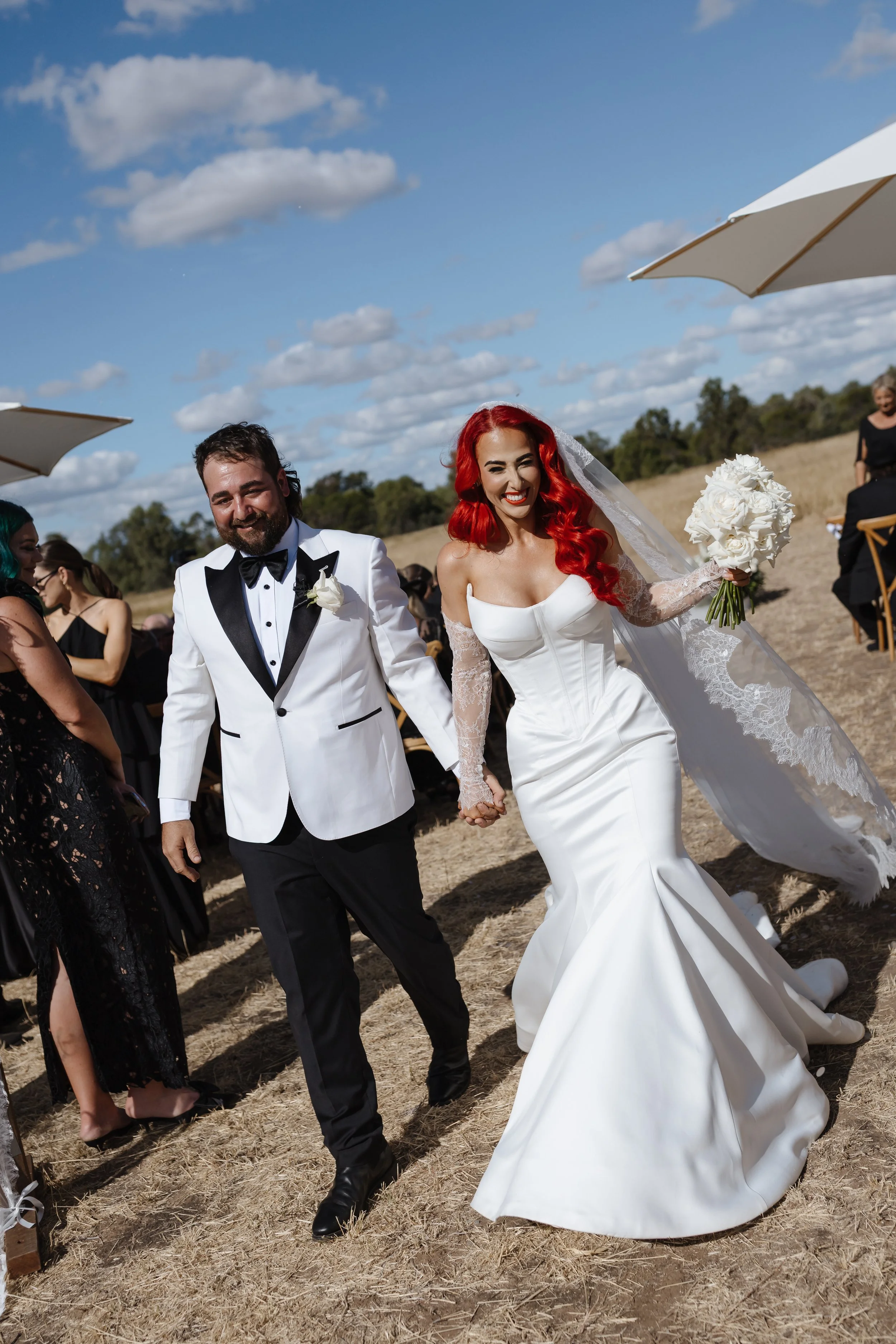 A couple in wedding attire walking outdoors on a sunny day with guests and umbrellas around them.