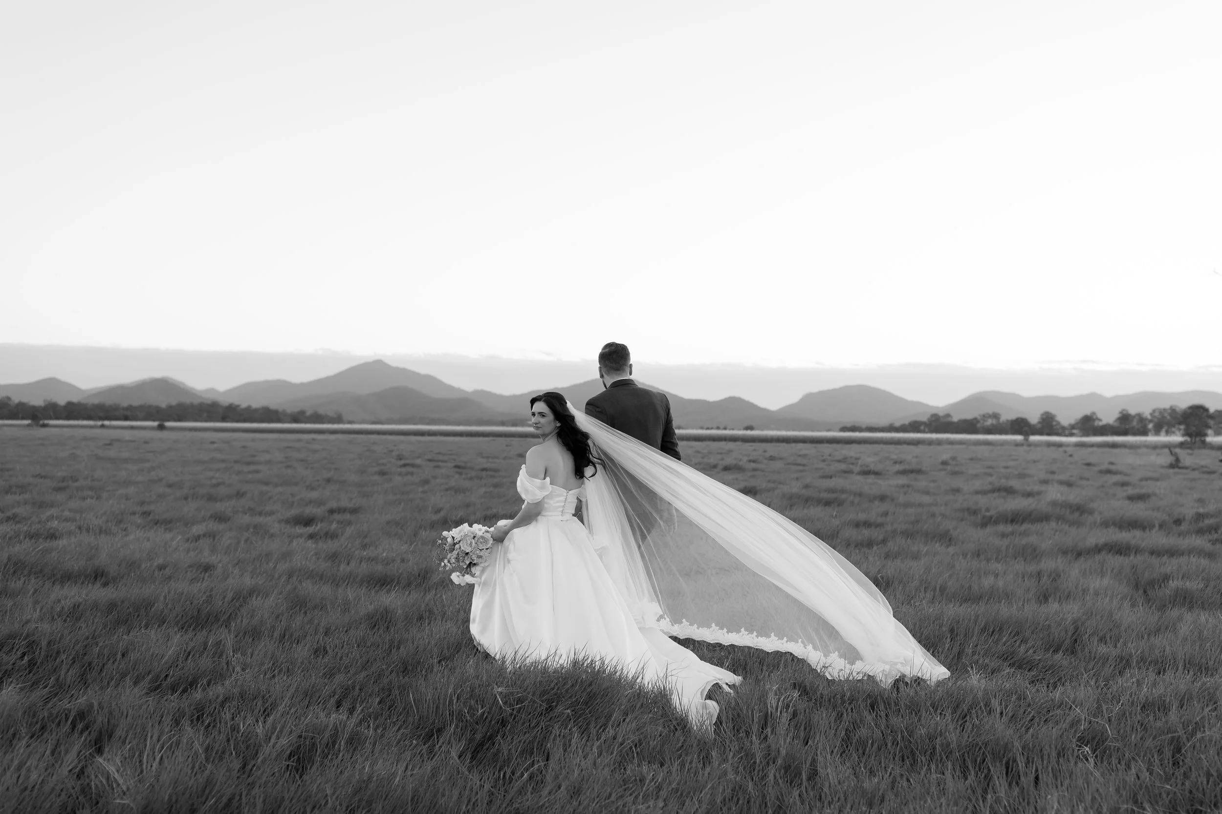 A black-and-white photo of a bride and groom standing in a grassy field, with mountains in the background. The bride is sitting, holding a bouquet, and smiling while looking to the side. The groom stands behind her, facing away, wearing a suit. The b