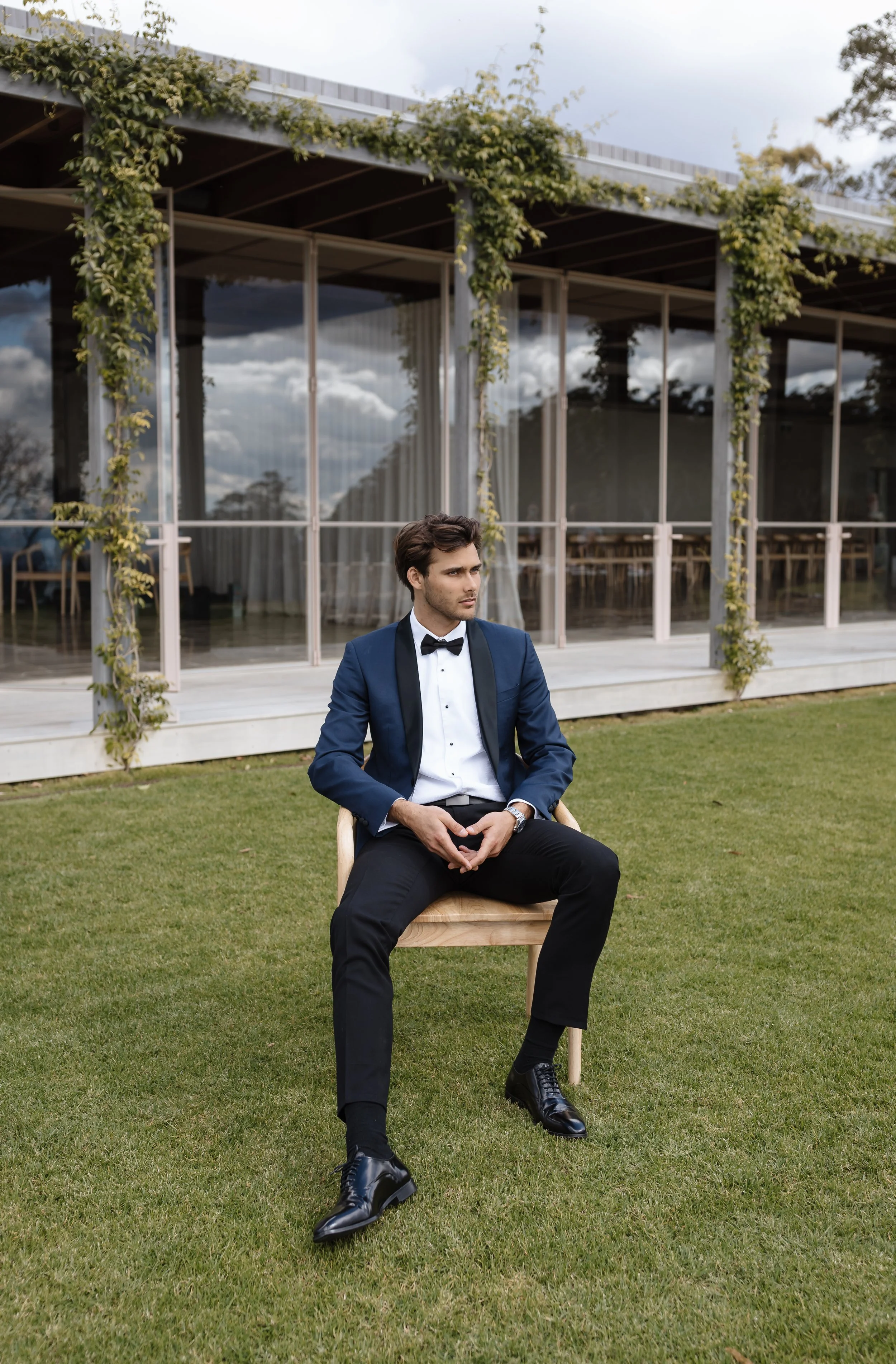 A man in formal attire, wearing a dark blue tuxedo and bow tie, sitting on a wooden chair outdoors on a grassy area in front of a modern glass building with ivy on the roof, looking to the right.