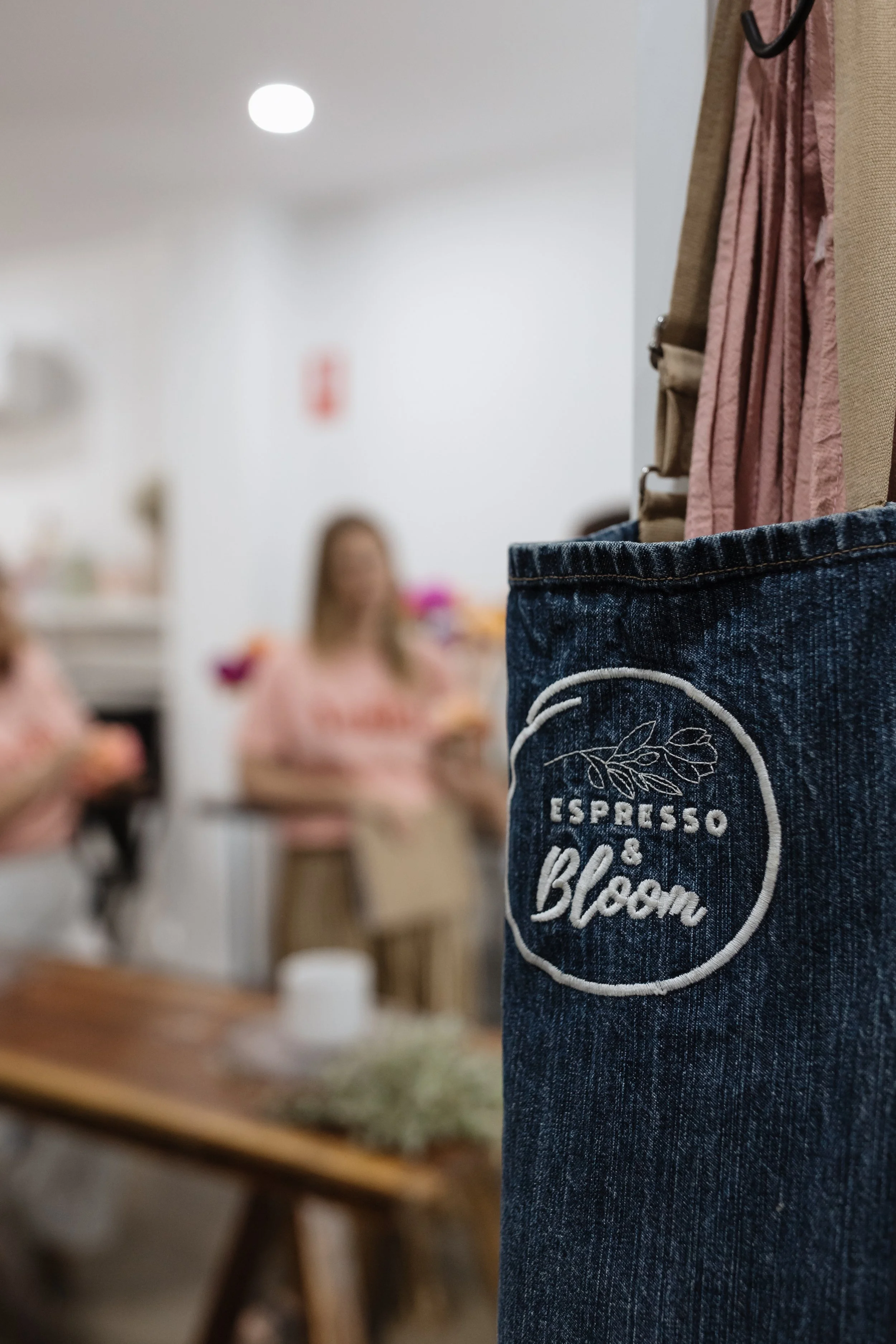 Close-up of a denim apron or shirt pocket with embroidery that reads "Espresso, I Love You" and a leaf design. In the background, blurred women in a room, some sitting at a table.