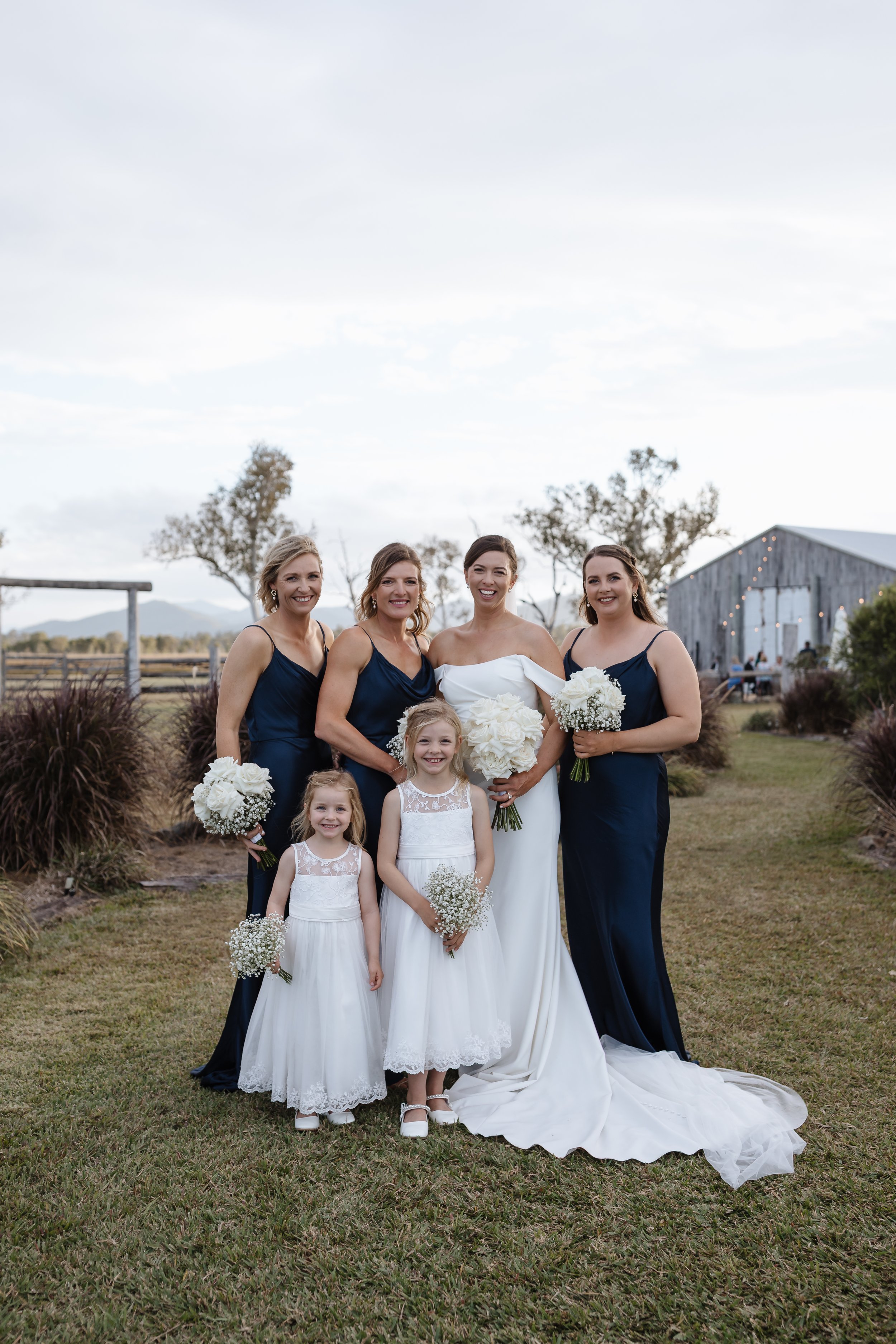 Group of women and young girls, dressed in formal attire, standing outdoors on a grassy area with trees and a barn in the background, smiling for a photo.