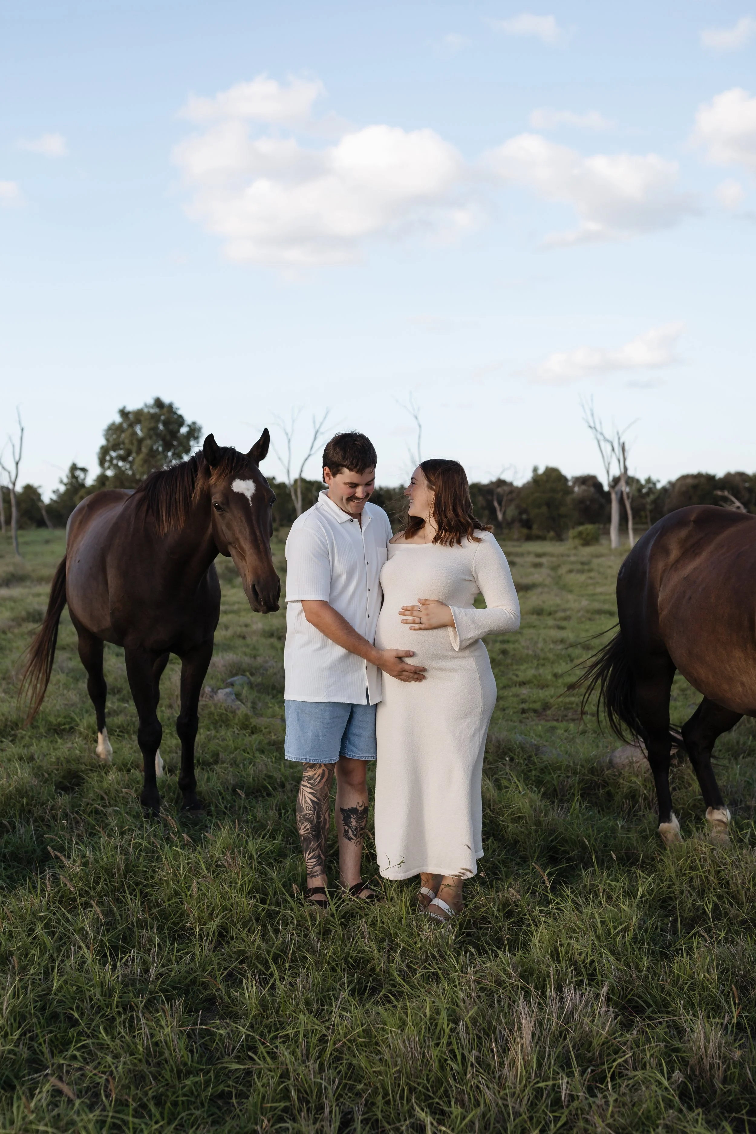 A pregnant woman in a white dress and a man with tattoos on his legs and arms stand together outdoors, smiling and looking at each other, with two horses nearby in a grassy field under a blue sky with some clouds.