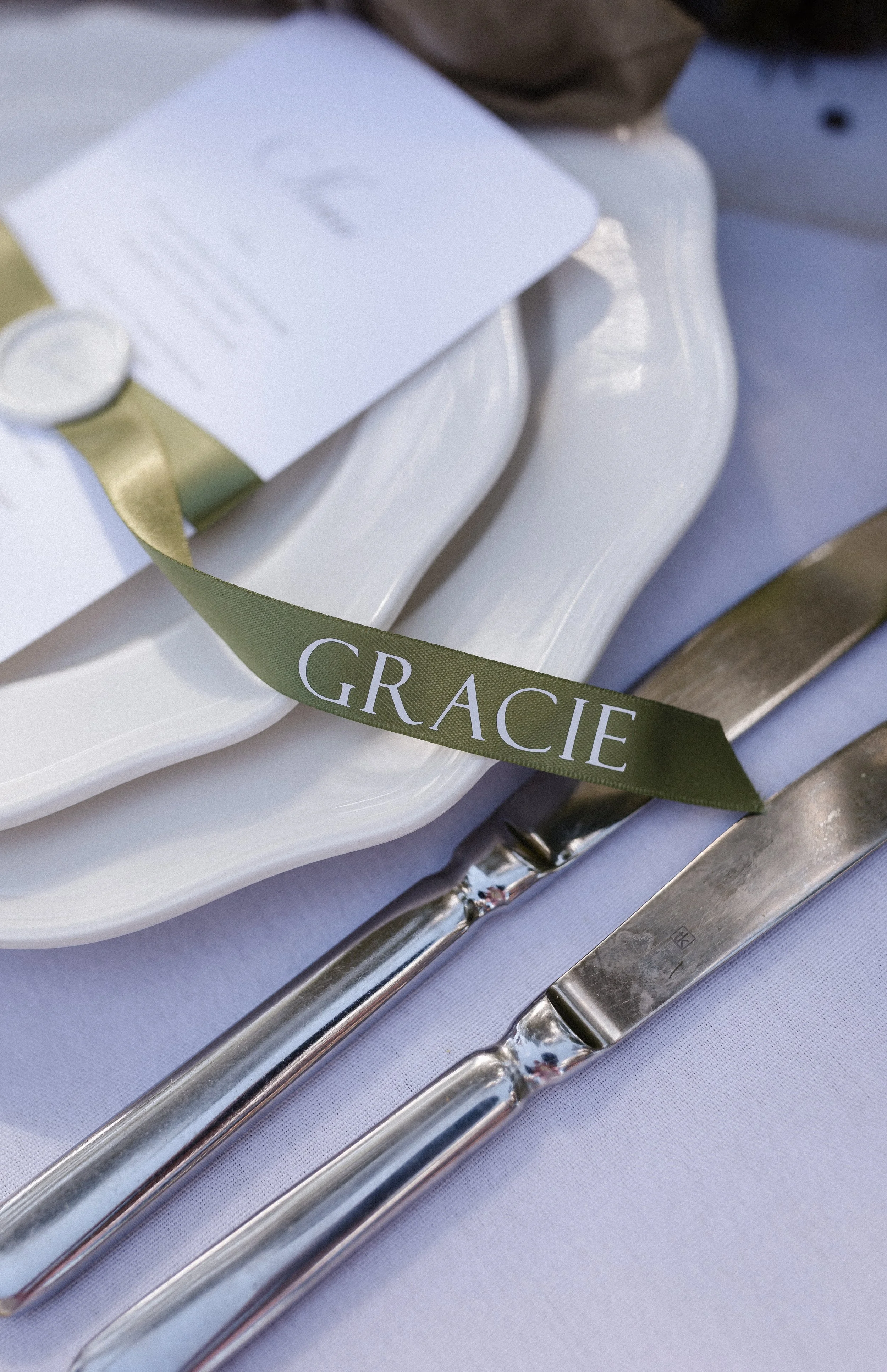 Close-up of a table setting with a white plate, a green ribbon with 'GRACIE' written on it, and silver utensils on a white tablecloth, with a blurred background.