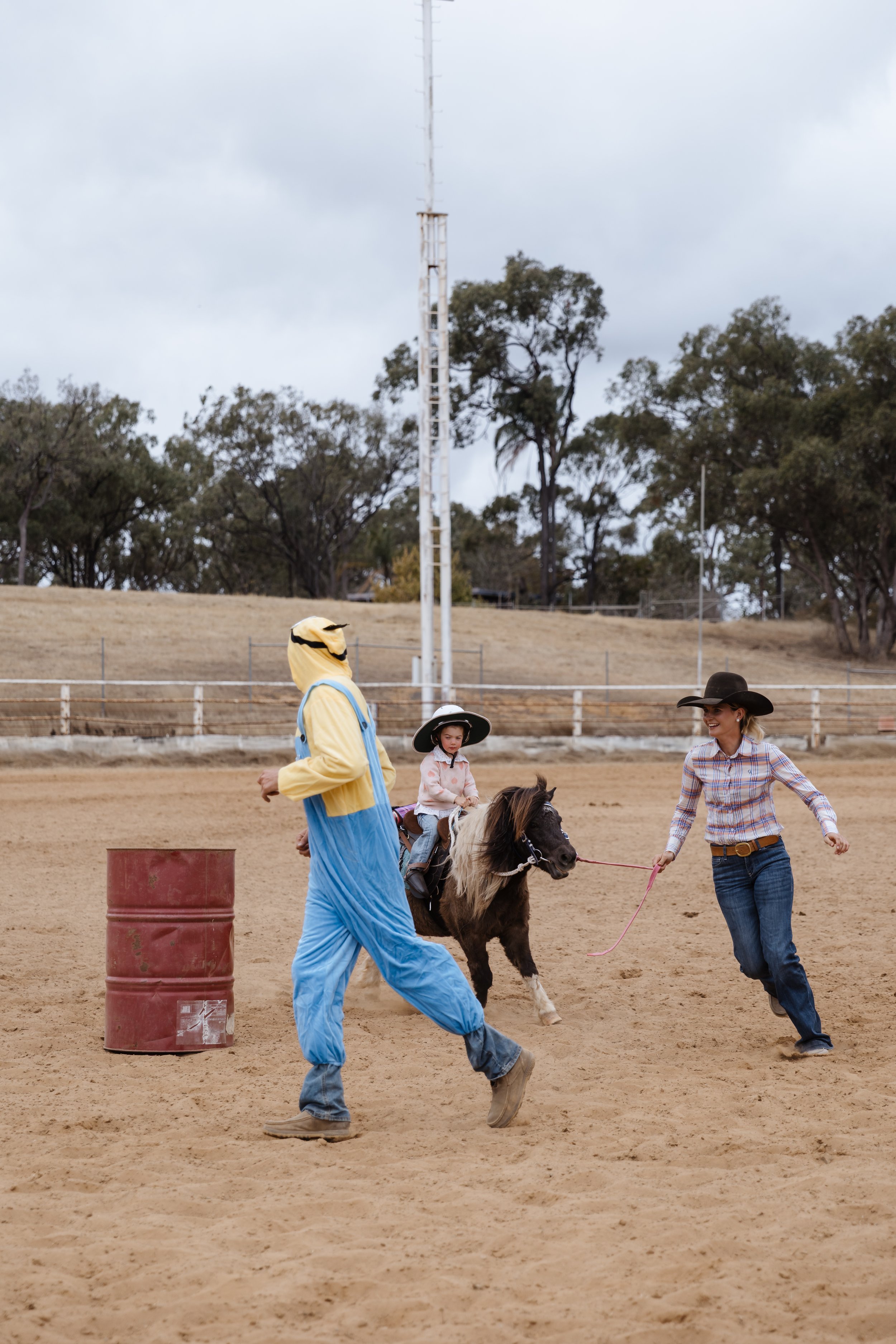 People riding a small horse in an outdoor rodeo or horse-riding event on a dirt field, with some participants wearing cowboy hats and casual clothing, and a rusted barrel and a fence in the background.