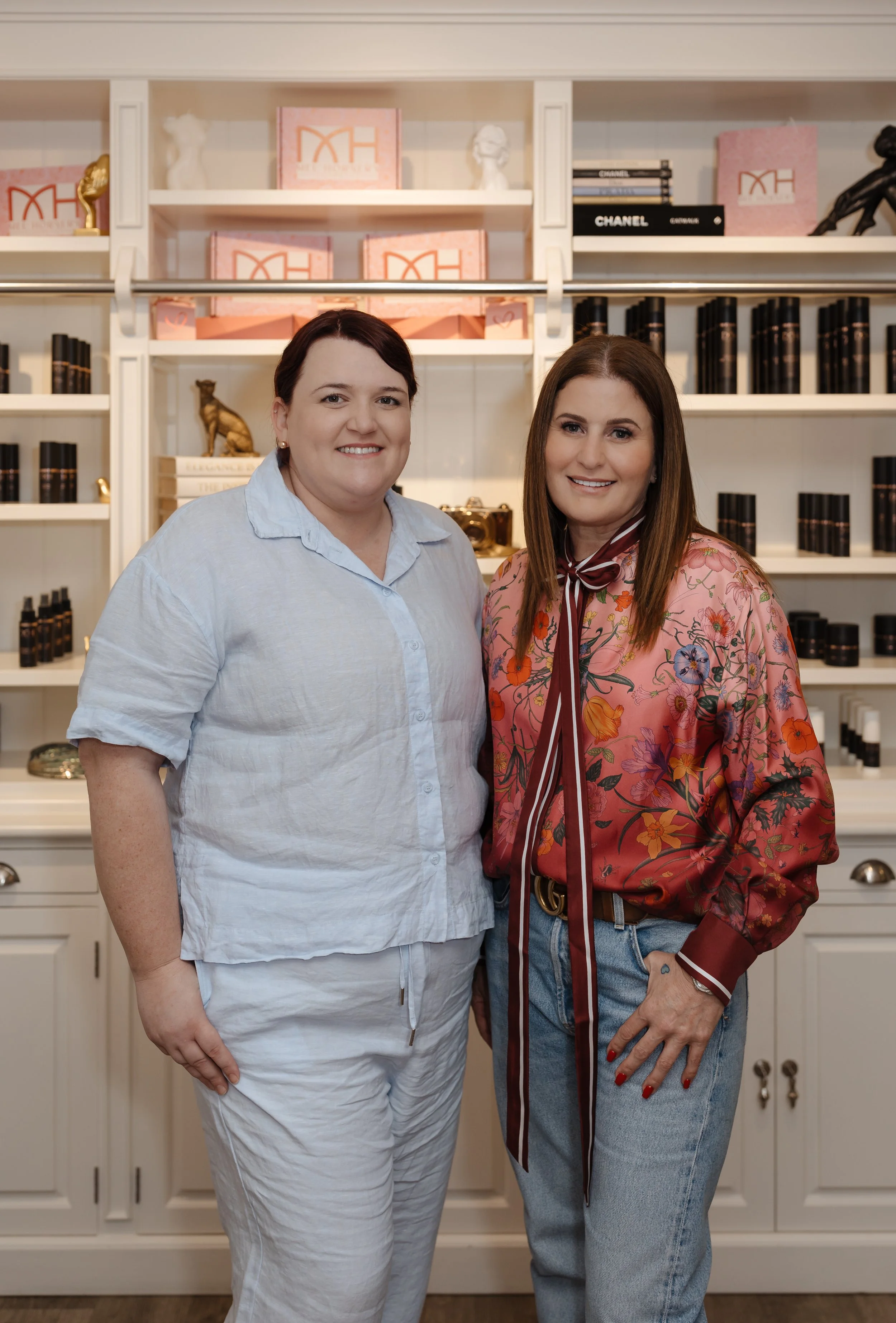 Two women smiling indoors in front of white shelves filled with books, decorative items, and pink boxes.