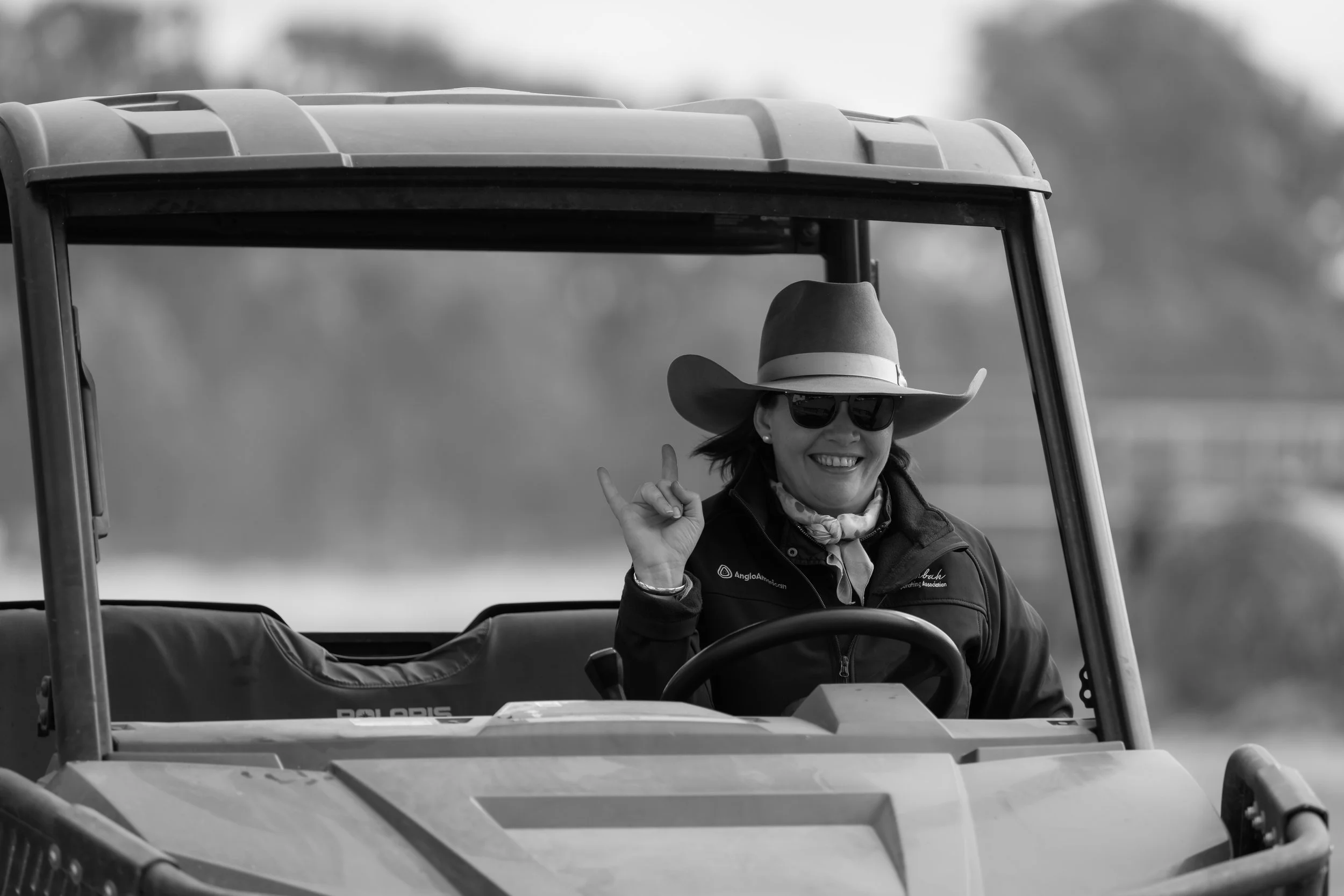 A woman wearing sunglasses, a wide-brimmed hat, and a jacket, smiling and making a peace sign while sitting in a golf cart.
