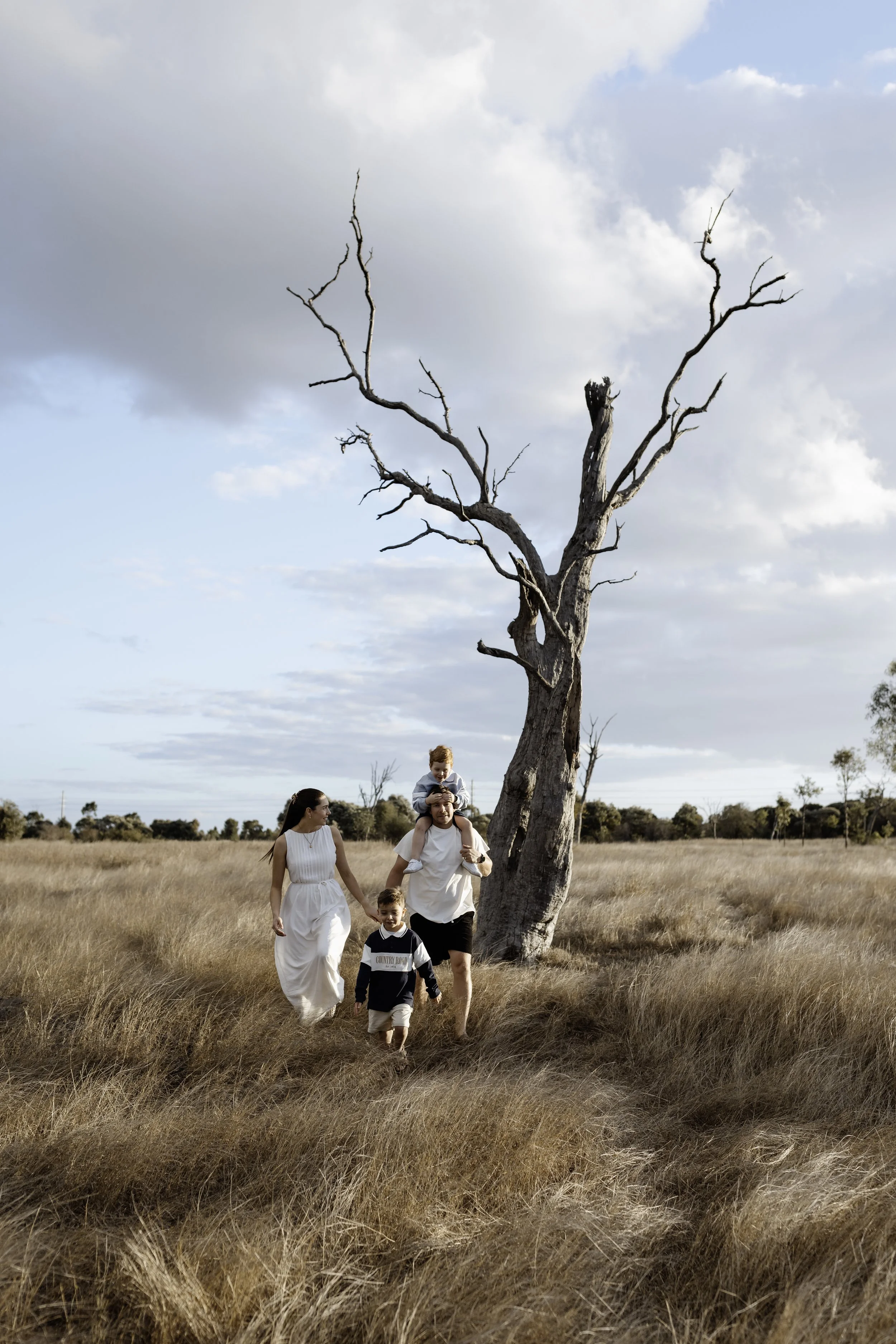 A family of five walking through a field of tall grass near a large, leafless tree under a partly cloudy sky.