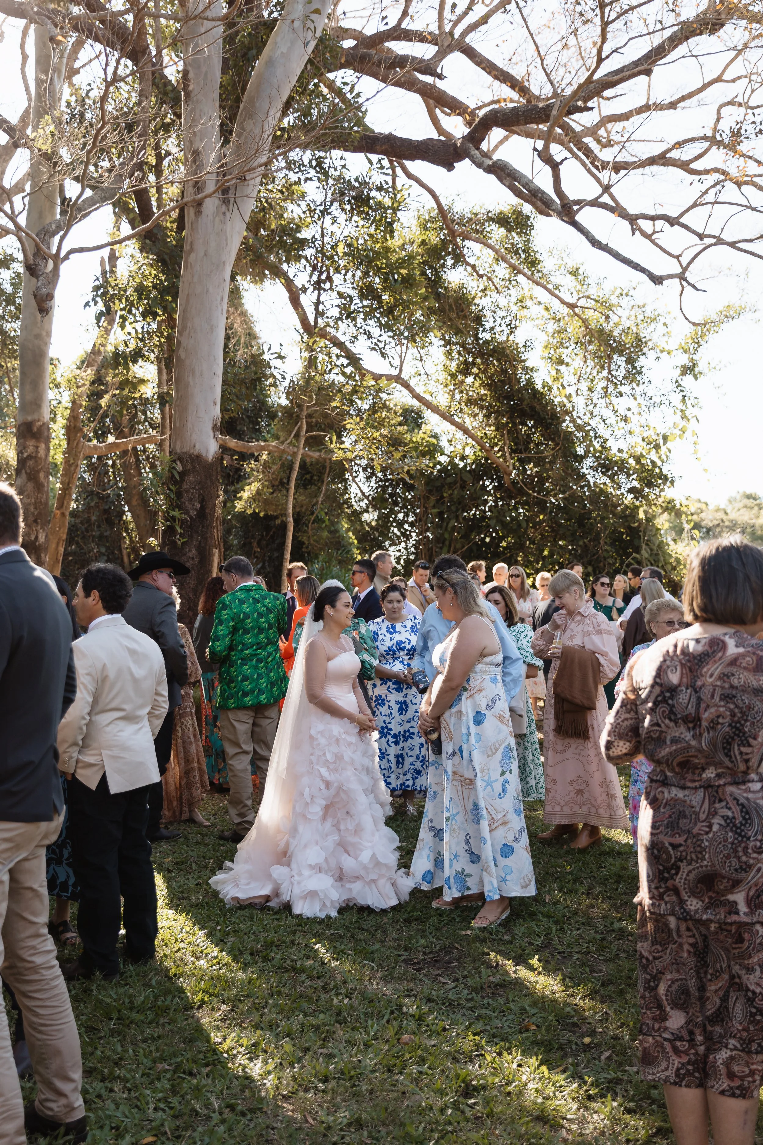 A group of people gathered outdoors, dressed in formal and semi-formal attire, standing on grass under large trees with bare branches and some green foliage. The focus is on a bride in a white wedding gown with ruffled layers, surrounded by guests, s