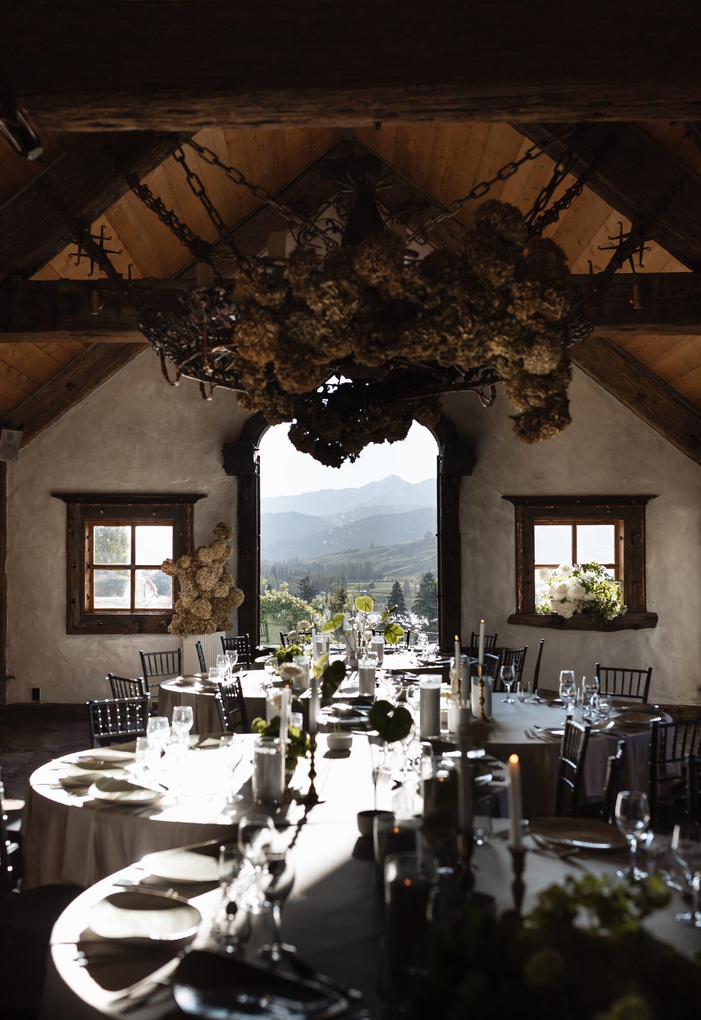 Upside-down image of a rustic wedding or event reception dining room with round tables set for dinner, large window showing scenic mountains and trees, and a chandelier hanging from a wooden ceiling.
