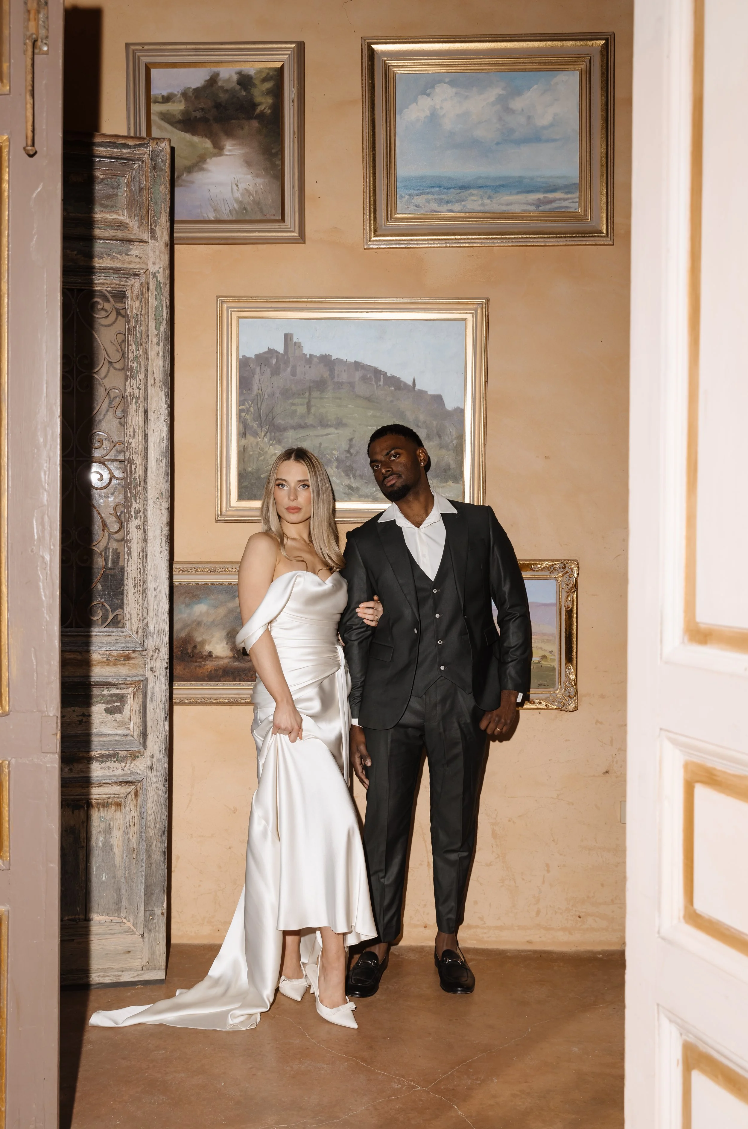 A woman in a white satin gown and a man in a black suit pose together in an art gallery. The woman has long blonde hair and is holding a clutch purse, while the man has short dark hair and a beard. The background features framed landscape paintings o