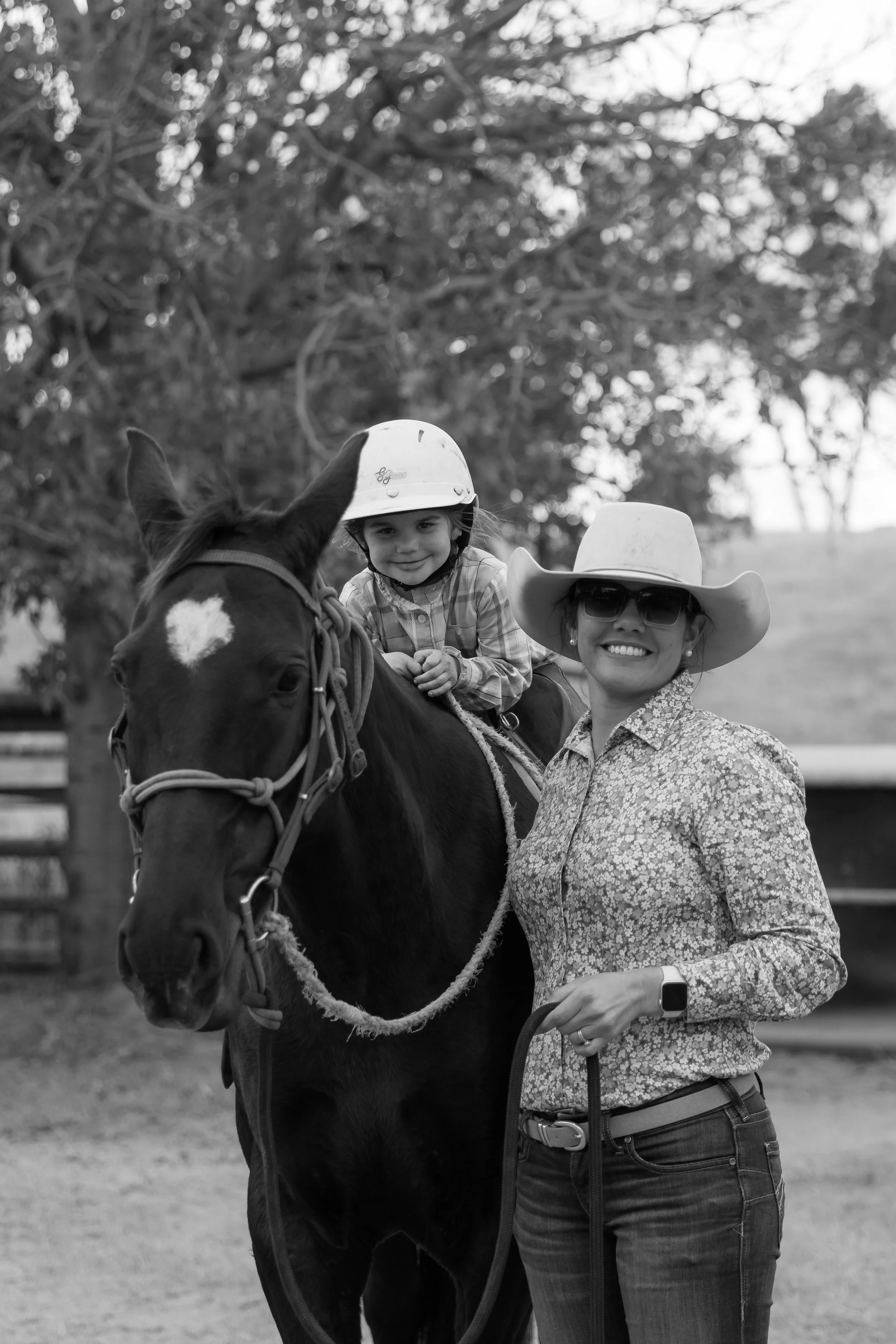 A woman wearing sunglasses and a wide-brimmed hat stands next to a horse, with a young girl riding the horse, both wearing helmets, outdoors with trees and a fence in the background.