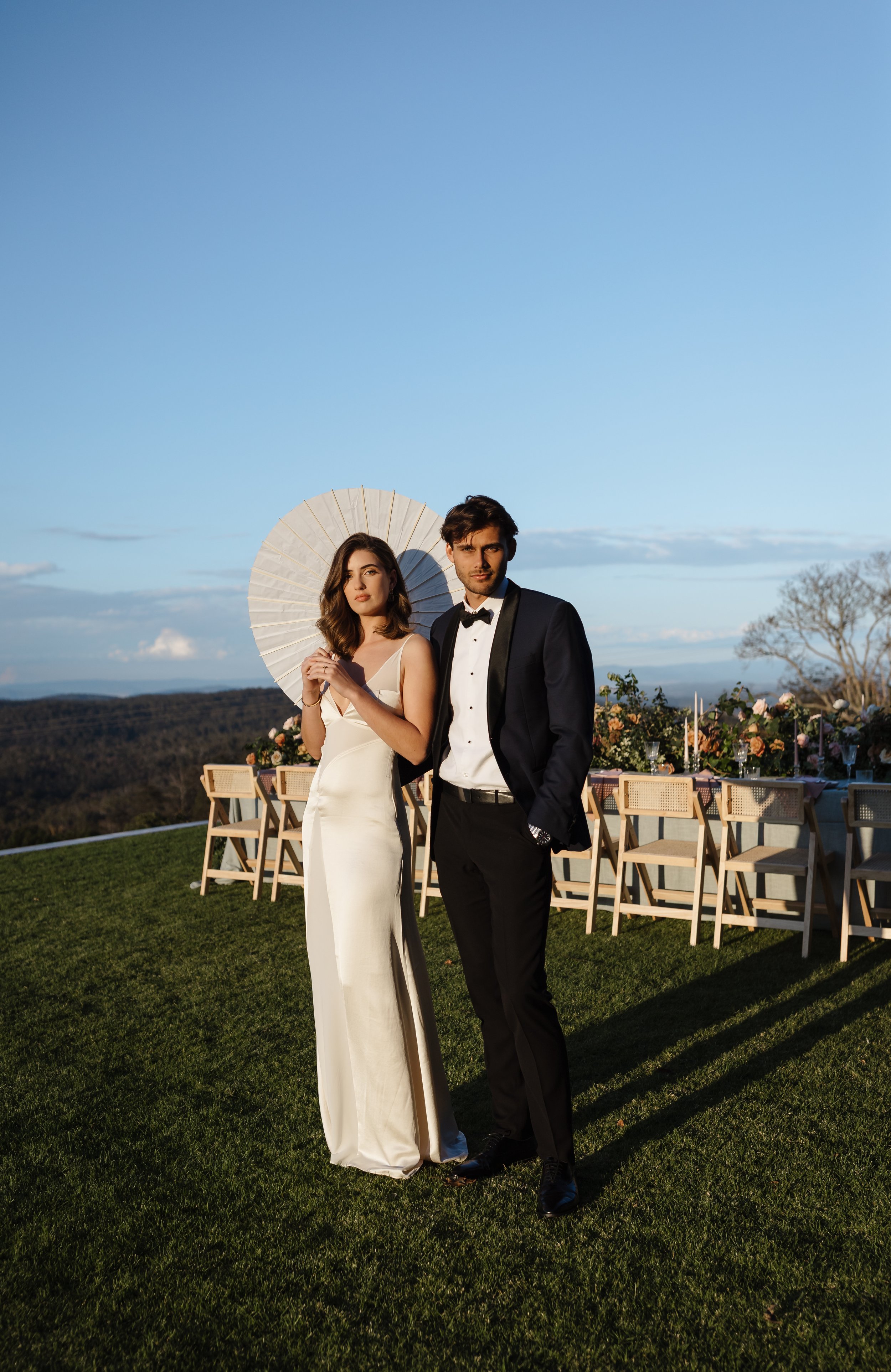 A couple dressed in formal wedding attire standing outdoors on a grassy area, with tables and chairs behind them, and mountains in the distance during sunset.