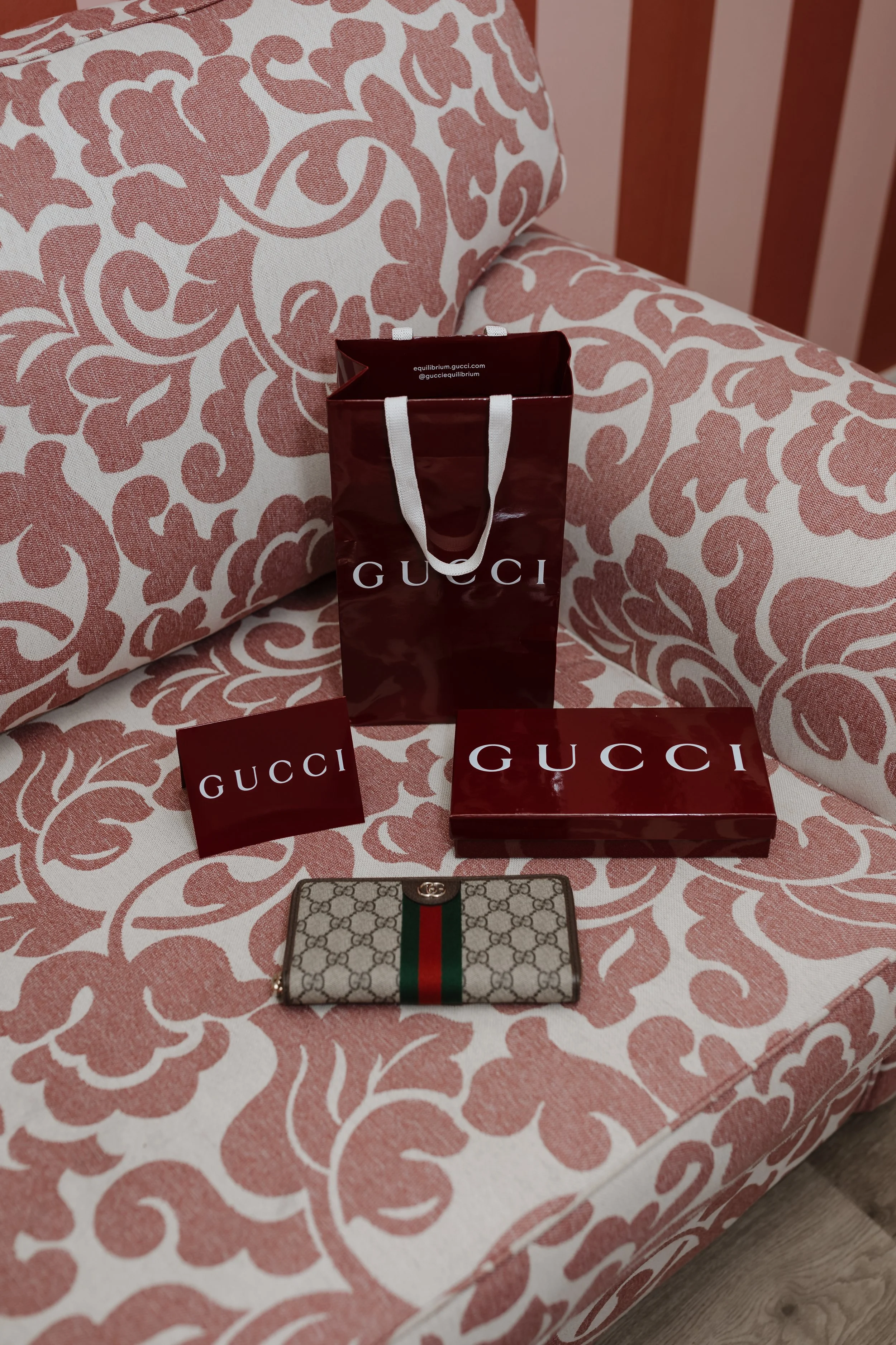 A pink and white patterned bedsheet with Gucci shopping bags and a wallet, alongside a small Gucci patterned wallet with a red and green stripe, on a wooden floor.