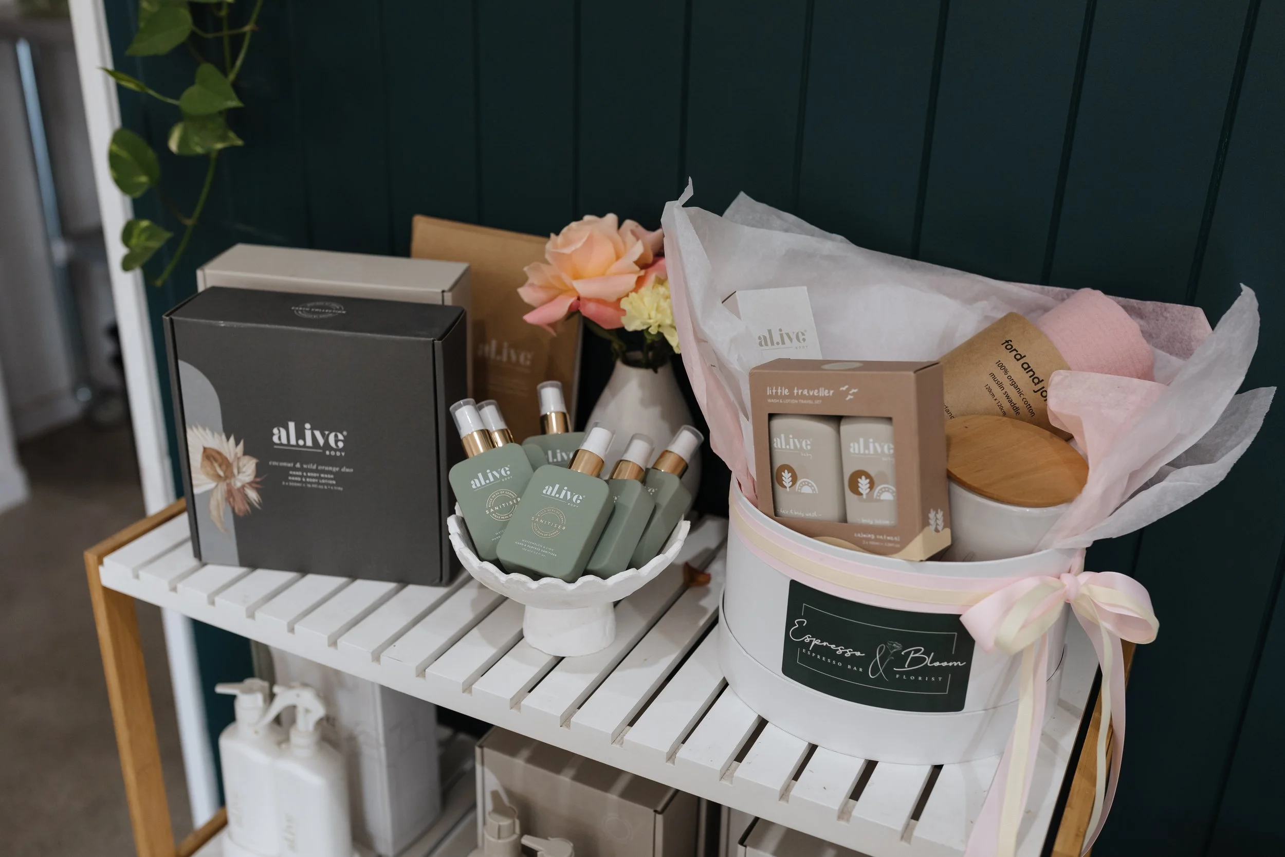 A display of bath and body products on a white slatted shelf, including black and white boxes, small bottles, and a bouquet of pink and cream flowers wrapped in tissue paper, in front of a dark green paneled wall.