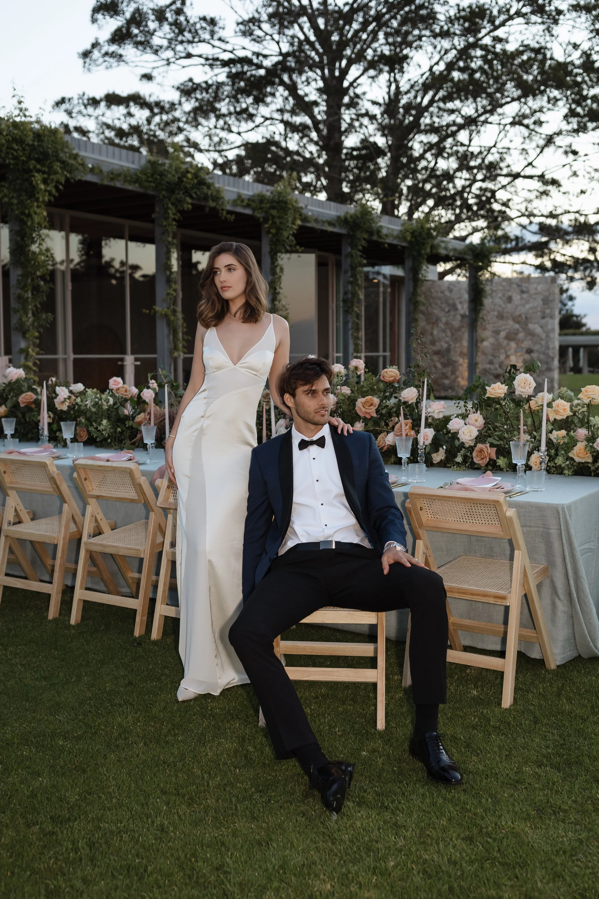 A woman in a white dress standing next to a seated man in a tuxedo at an outdoor wedding reception with decorated tables and woodland scenery in the background.