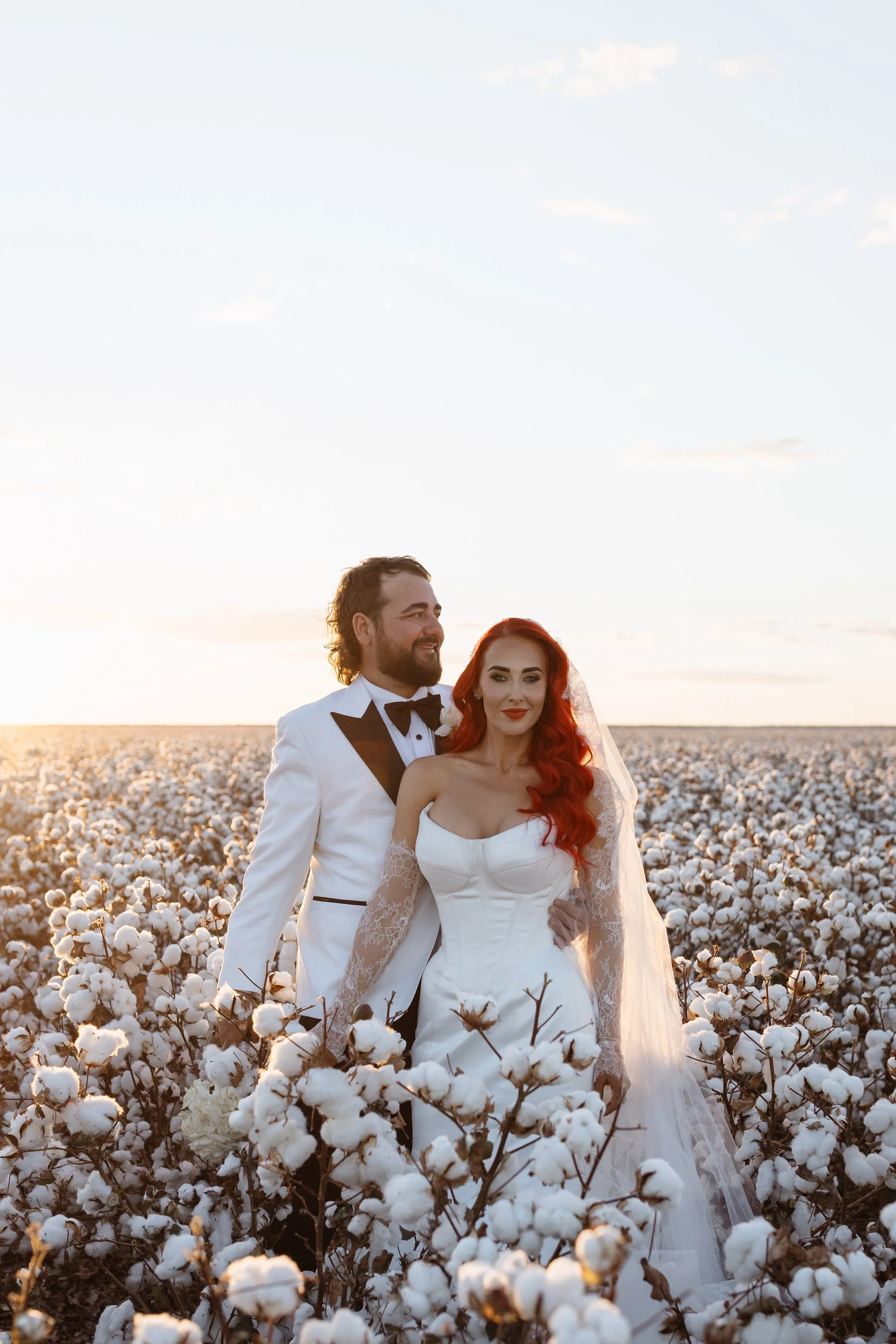 A bride and groom standing in a cotton field at sunset, the groom in a white tuxedo with black lapels and bowtie, the bride in a strapless white wedding dress with lace sleeves and a long train, both smiling and looking in different directions.
